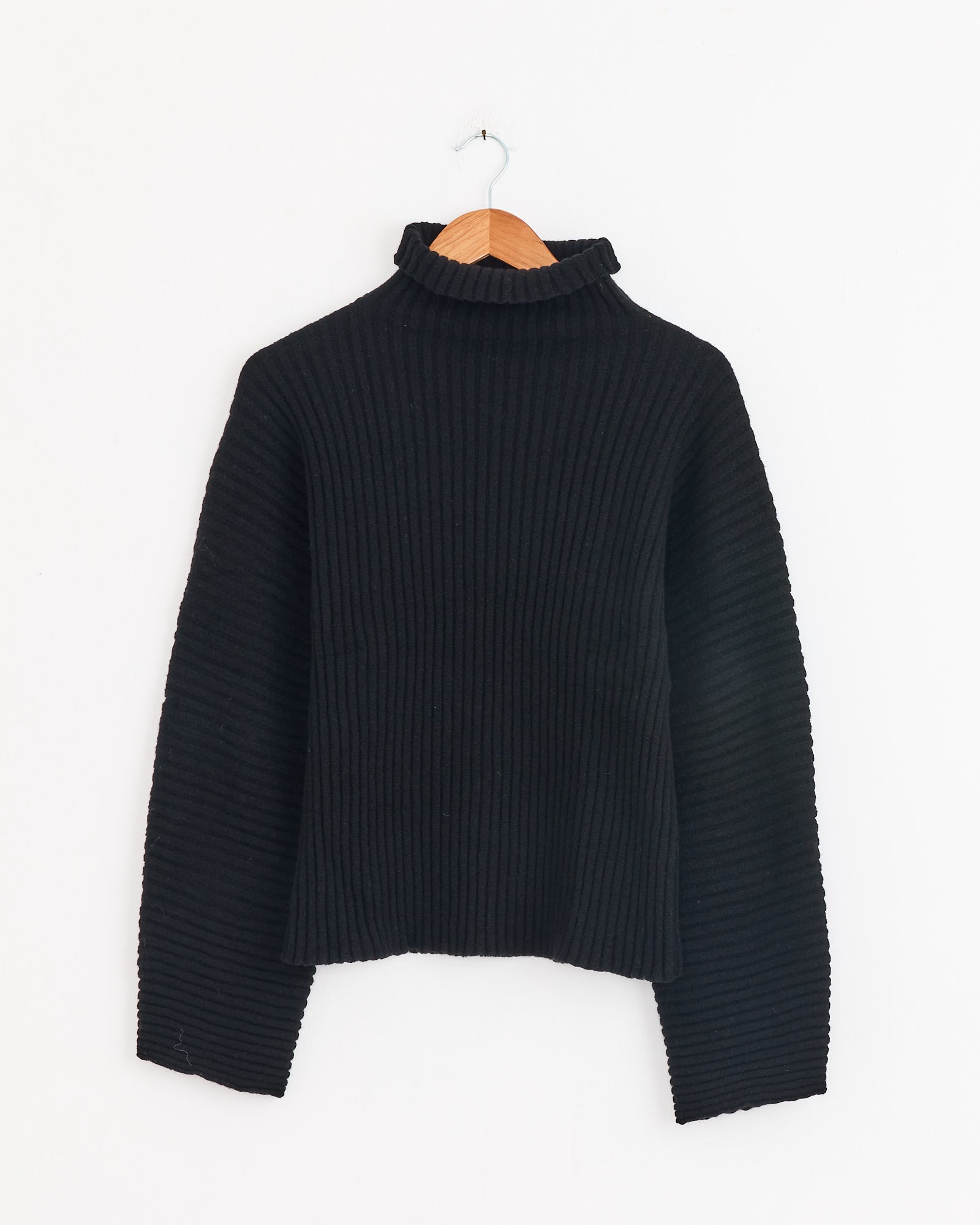 The Baserange Kai Turtleneck in Black hangs on a wooden hanger against a plain white wall.