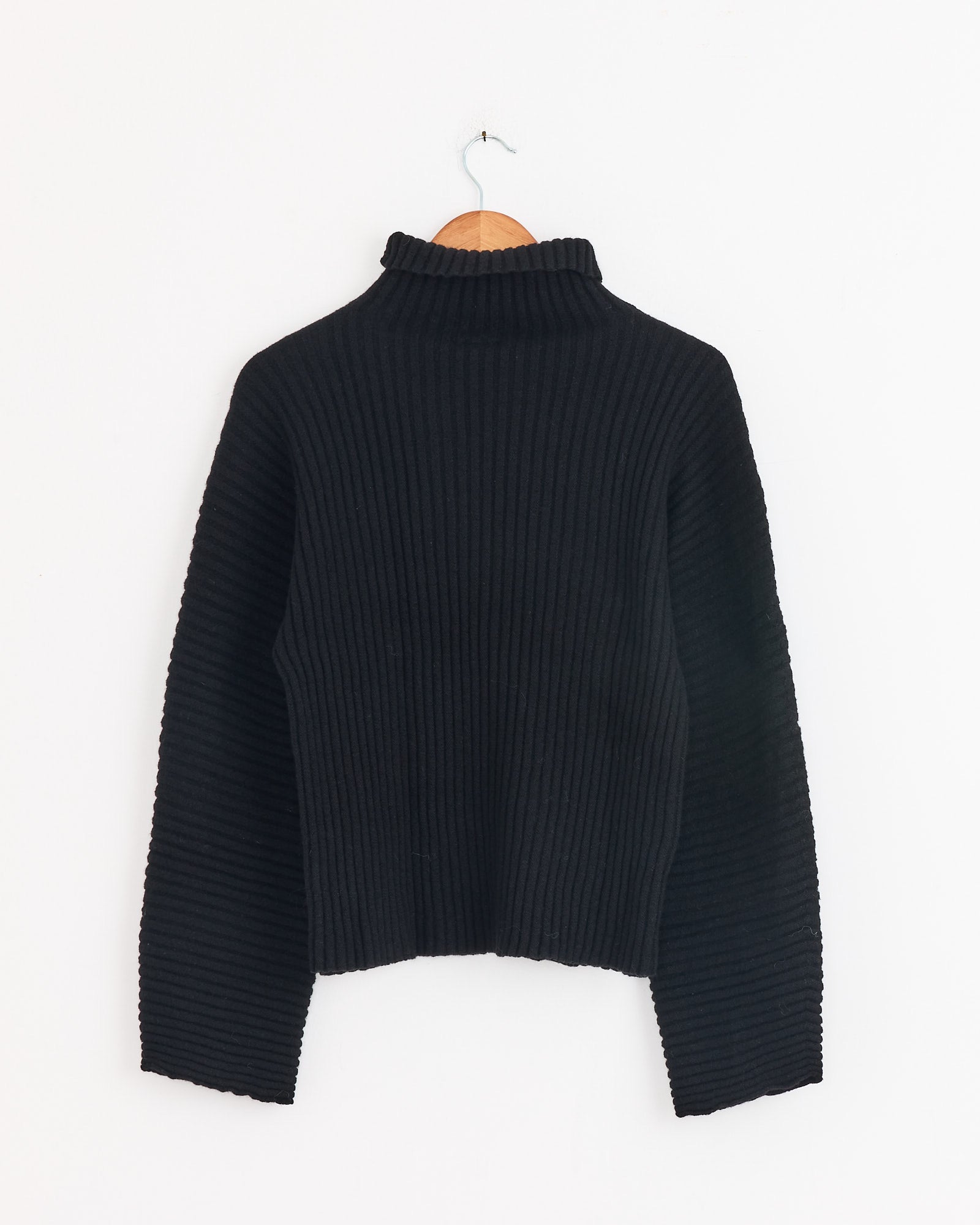 The Baserange Kai Turtleneck in Black, featuring long sleeves and ribbed fabric, hangs on a wooden hanger against a white wall, highlighting the signature style of the brand's collection.