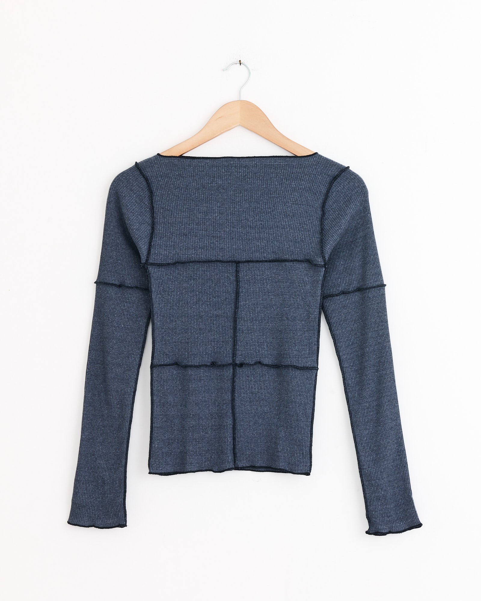 The Baserange Cinder Tee in Dark Denim is a blue long-sleeve ribbed top with black exposed seams and lettuce-edge hems, shown on a wooden hanger against a white background.