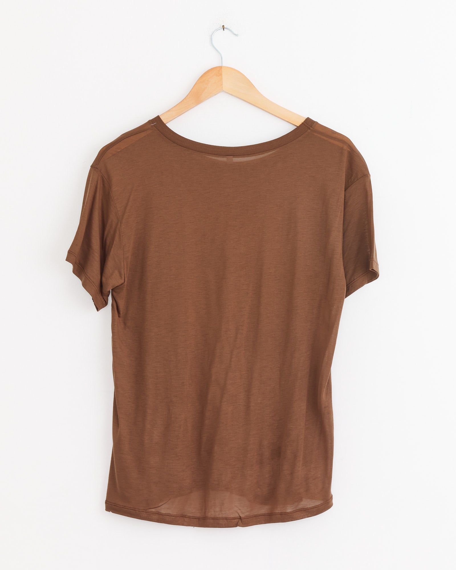 The Baserange Loose Tee in Ocular is shown from the back on a wooden hanger against a white wall, highlighting its relaxed silhouette and lightweight, casual design.