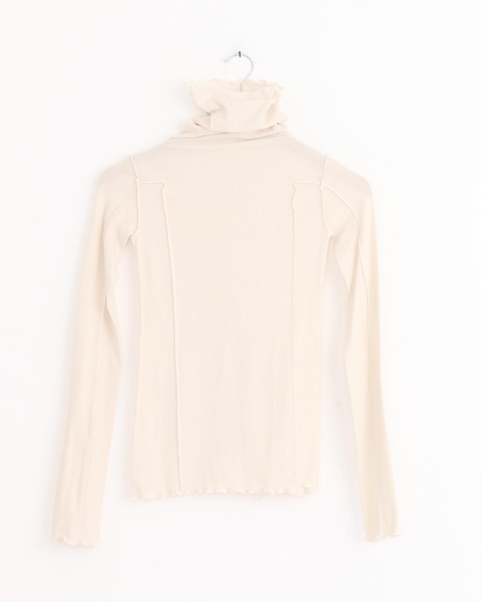 The Baserange Omato Turtleneck in Off White is an organic cotton top with long sleeves, a ruffled collar and hem, shown hanging on a hook against a white background.