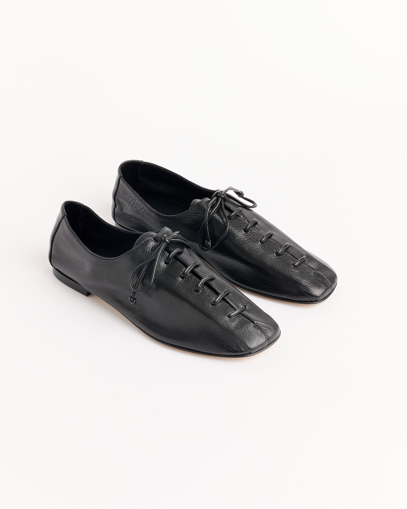 A pair of Hereu Plegada Supple Shiny Shoes in black, featuring a square toe, lace-up design, flat soles, and minimalist stitching, displayed on a white background. For fit or sizing questions, contact our online chat for help.