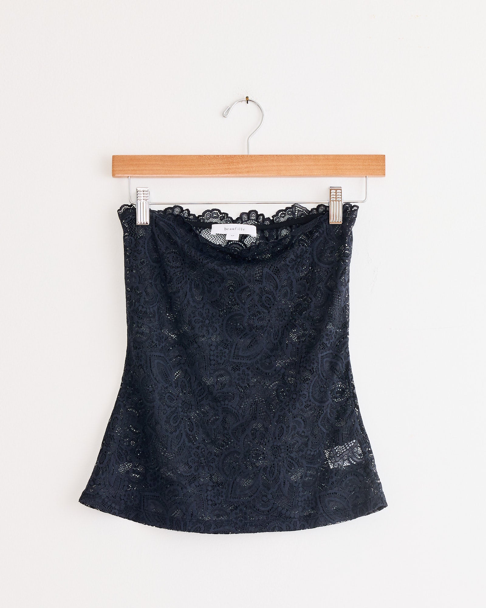 The Anise Blouse in Black by Beaufille, a strapless lace design with a subtle paisley pattern, is shown on a wooden hanger against a white background.