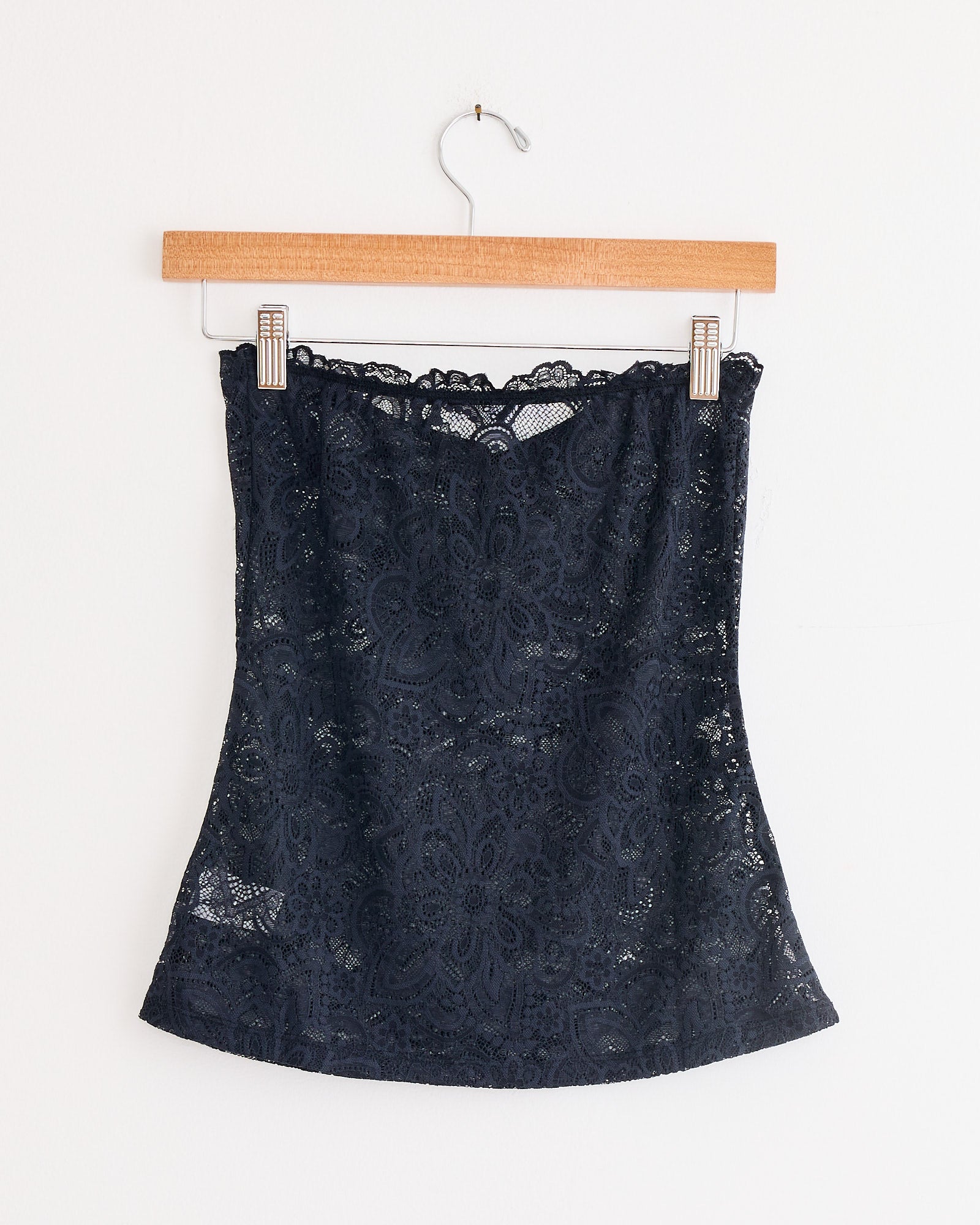 The Anise Blouse in Black by Beaufille, a strapless lace top with a floral pattern, hangs on a wooden hanger against a plain white wall.