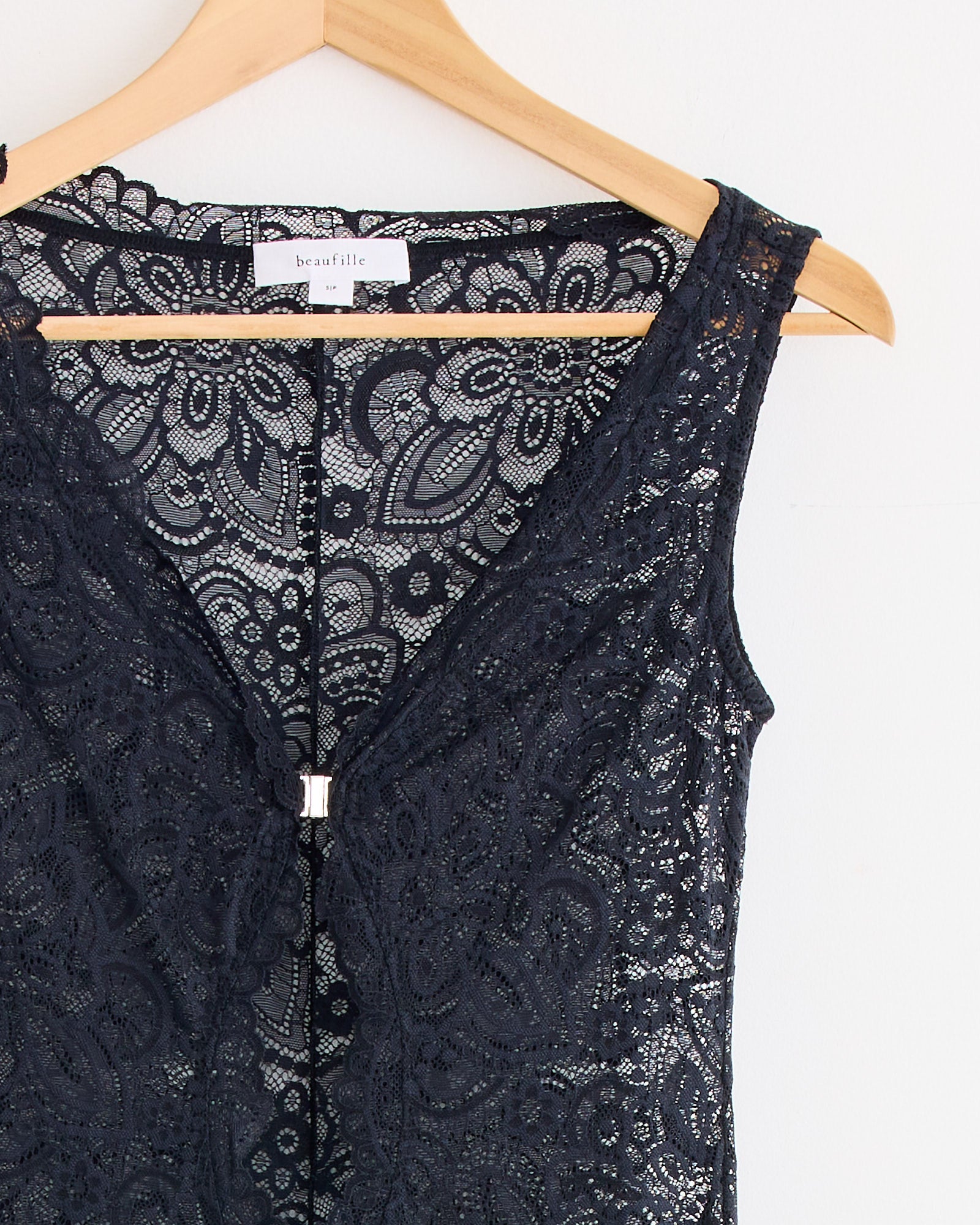 The Twain Lace Dress in Black by Beaufille is a slim fit, sleeveless lace dress with intricate floral patterns, a V-neckline, and a small central metal clasp, shown hanging on a wooden hanger against a white background.