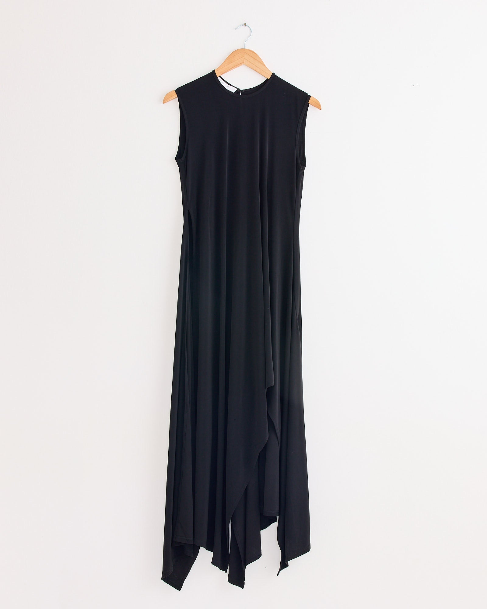 The Beaufille Hari Dress in Black, an elegant sleeveless piece with an asymmetrical draped hem, hangs on a wooden hanger against a plain white wall.