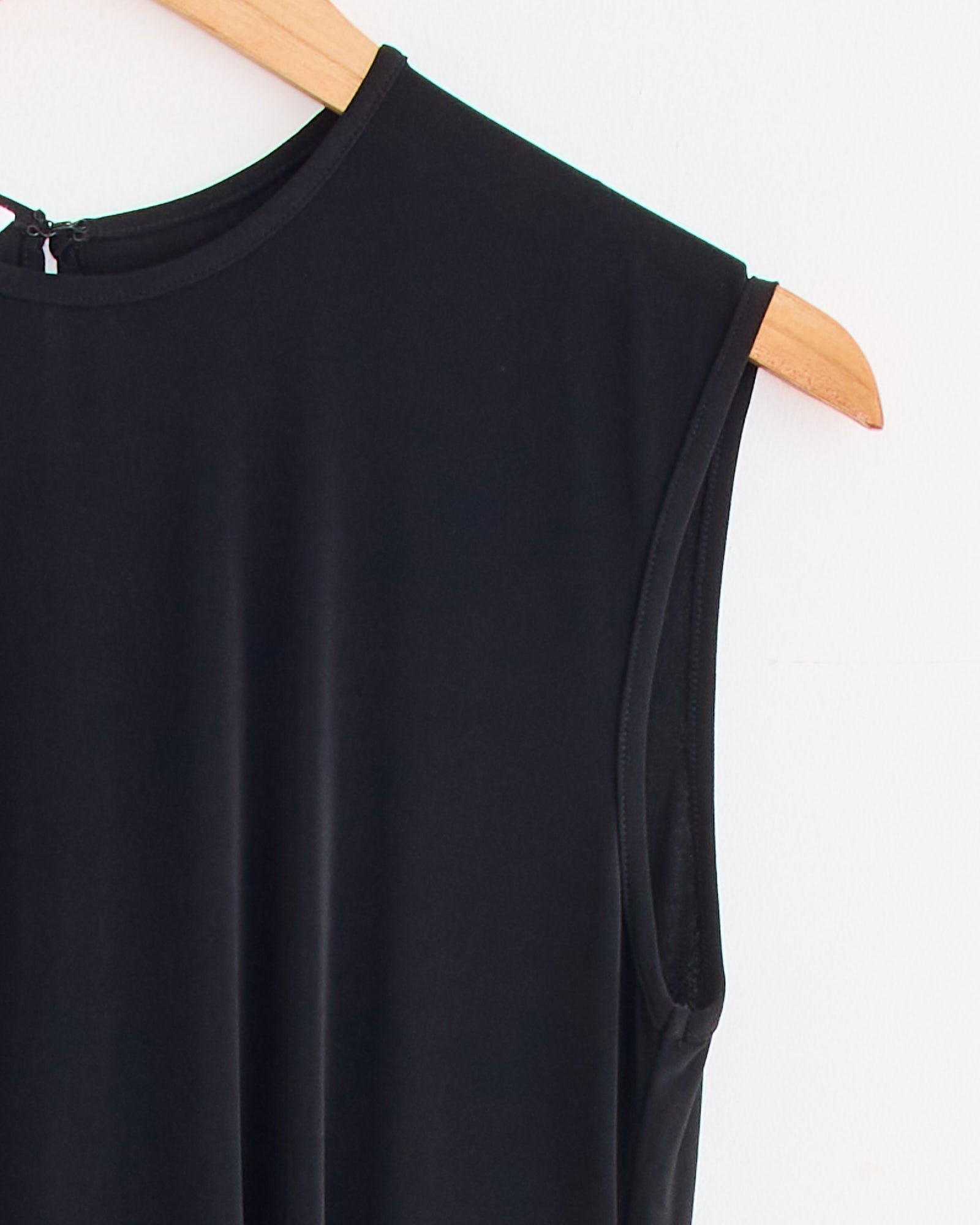 The Hari Dress in Black by Beaufille, featuring a sleeveless design, round neckline, and flattering waist tie, is displayed on a wooden hanger against a white background.