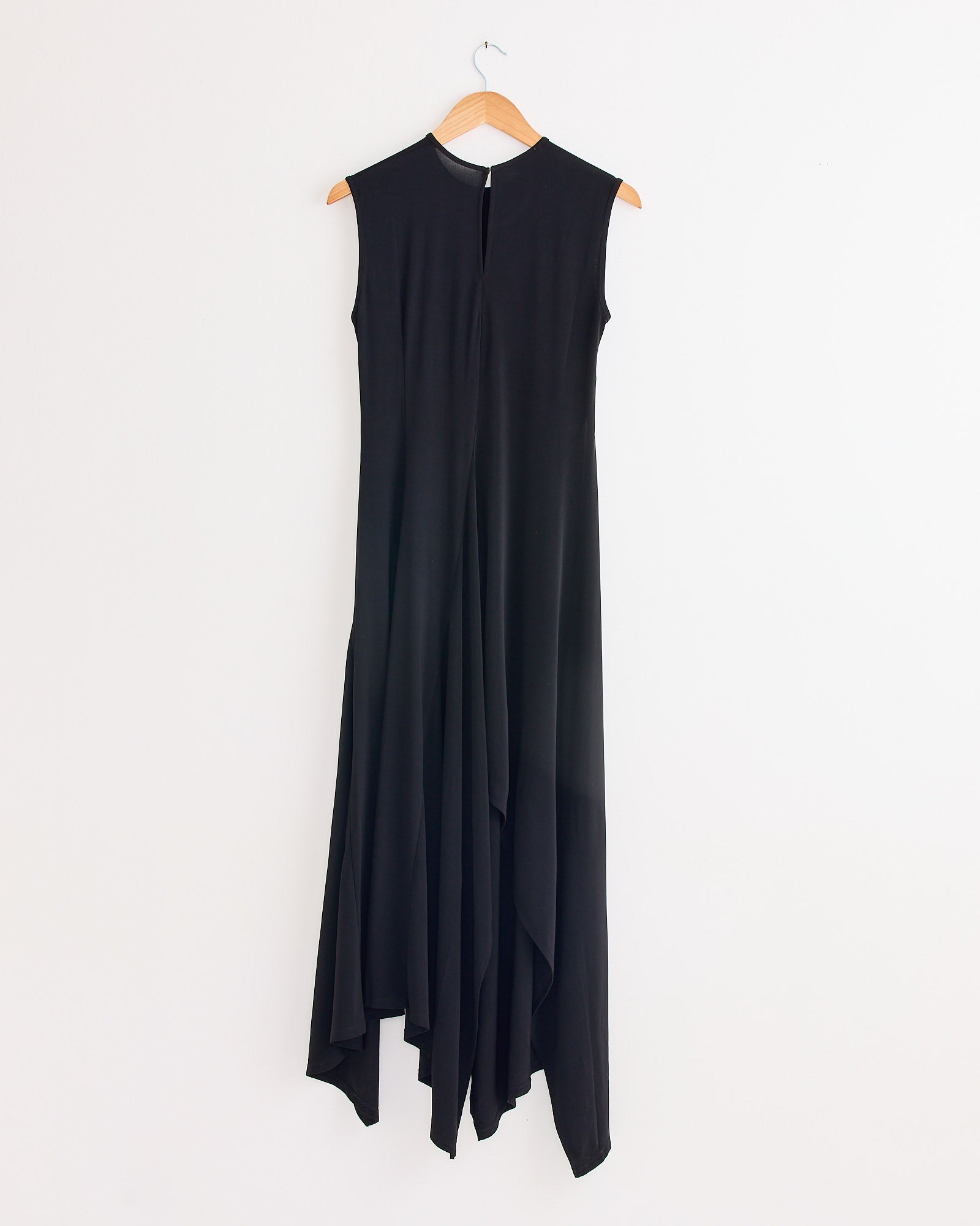 The Beaufille Hari Dress in Black, a sleek sleeveless piece with a high neckline, waist tie, and asymmetrical flowy hem, is displayed on a wooden hanger against a plain white wall.