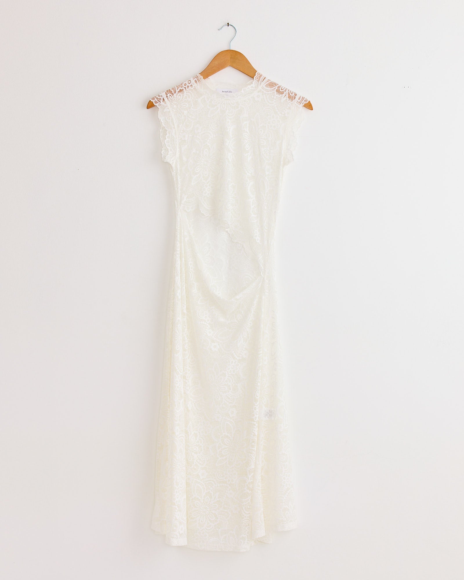 The Silva Dress in White by Beaufille is a white, short-sleeved lace crew neck dress with a fitted waist and flared skirt, displayed on a wooden hanger against a plain white wall.