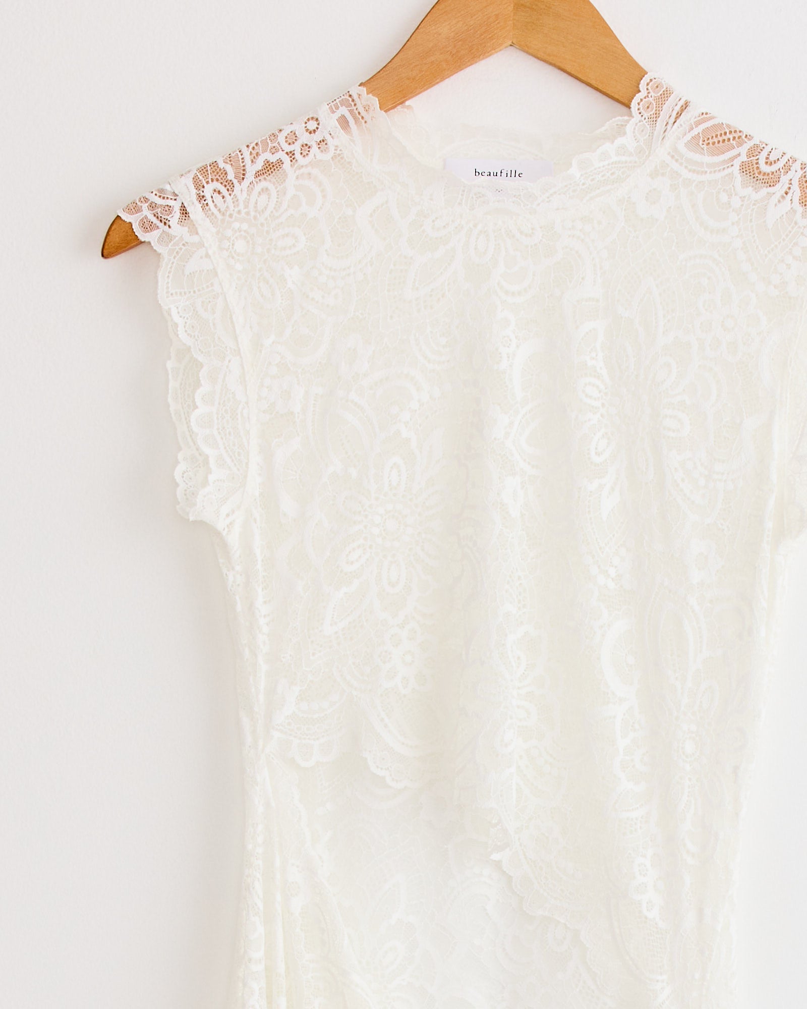 The Silva Dress in White by Beaufille hangs on a wooden hanger against a white wall, showcasing its floral-patterned lace fabric, scalloped edge, cap sleeves, crew neck, and intricate lace detailing throughout.