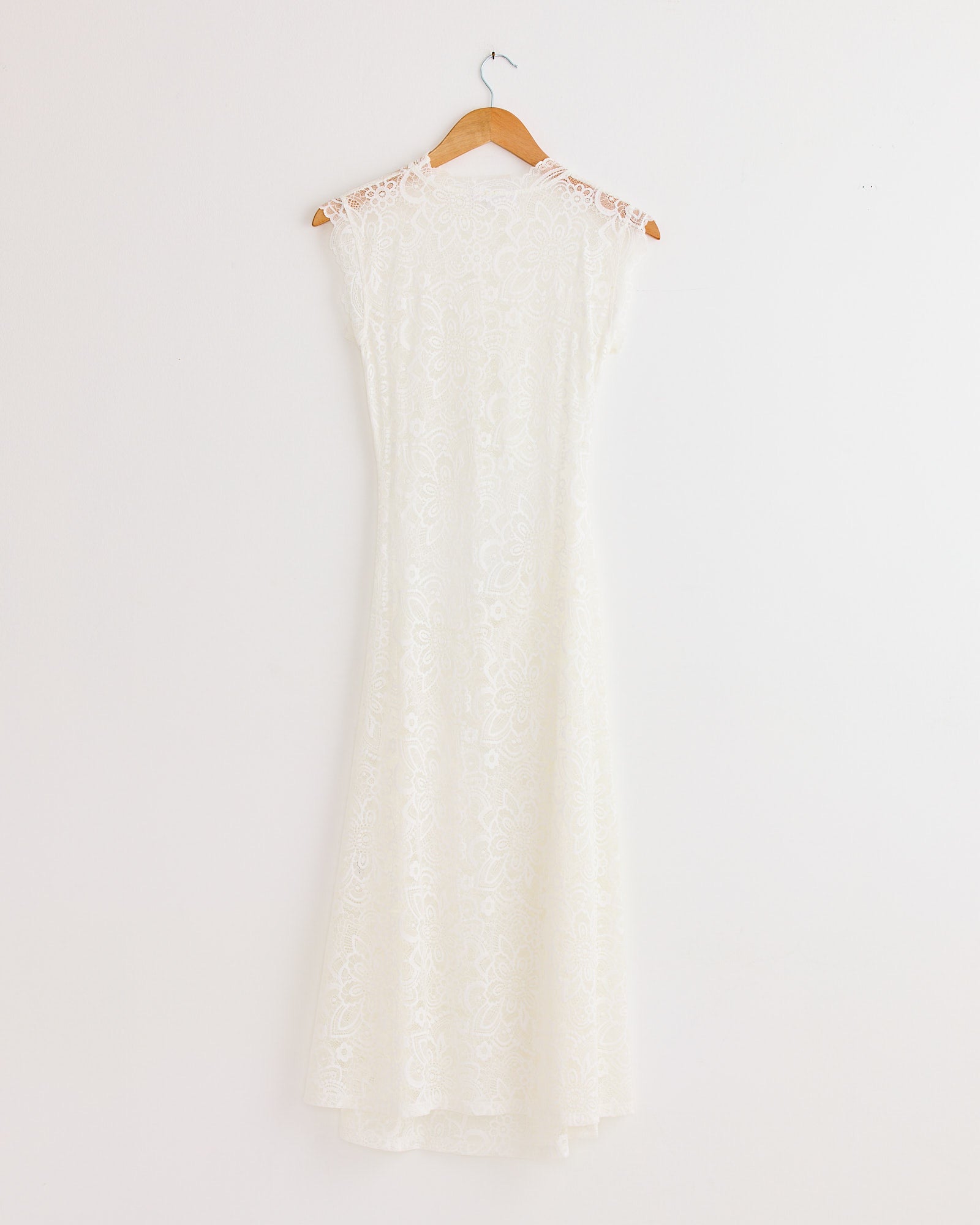 The Silva Dress in White by Beaufille is a sleeveless midi dress with delicate white lace and a subtle floral pattern, featuring a crew neck and flared skirt. Timelessly elegant, it hangs gracefully on a wooden hanger.