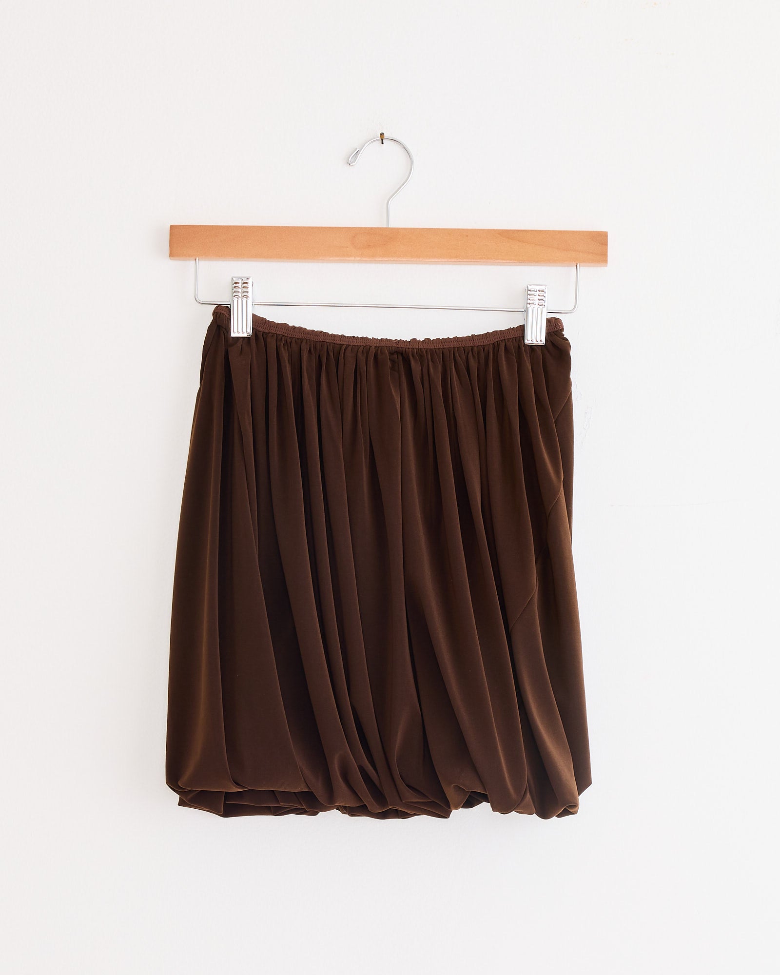 The Twisted Maro Skirt in Brown by Beaufille, featuring pleats and an elastic waist, hangs on a wooden hanger against a plain white wall.