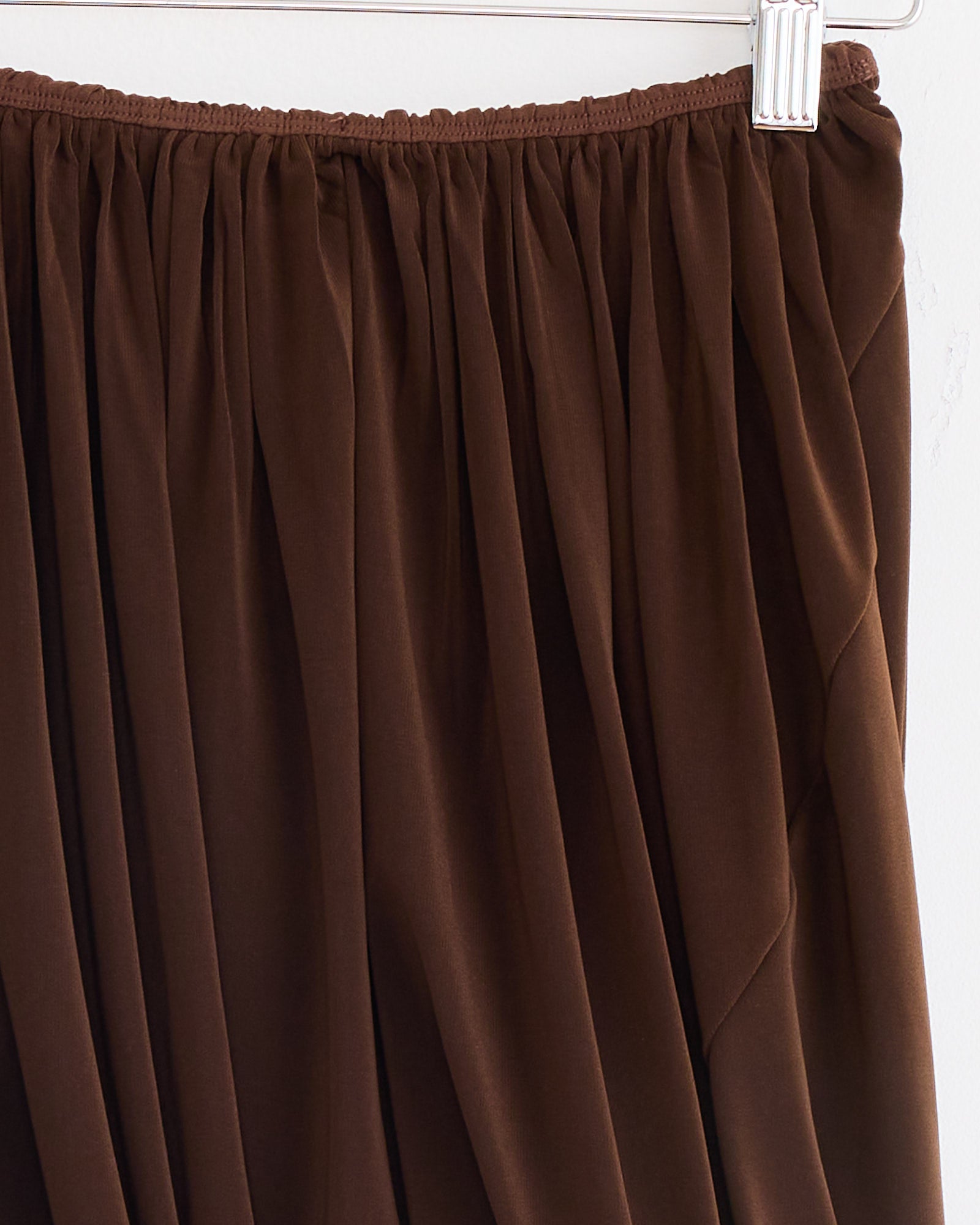 The Twisted Maro Skirt in Brown by Beaufille is a brown pleated jersey mini skirt with an elasticized waist, shown hanging on a white hanger against a white background.