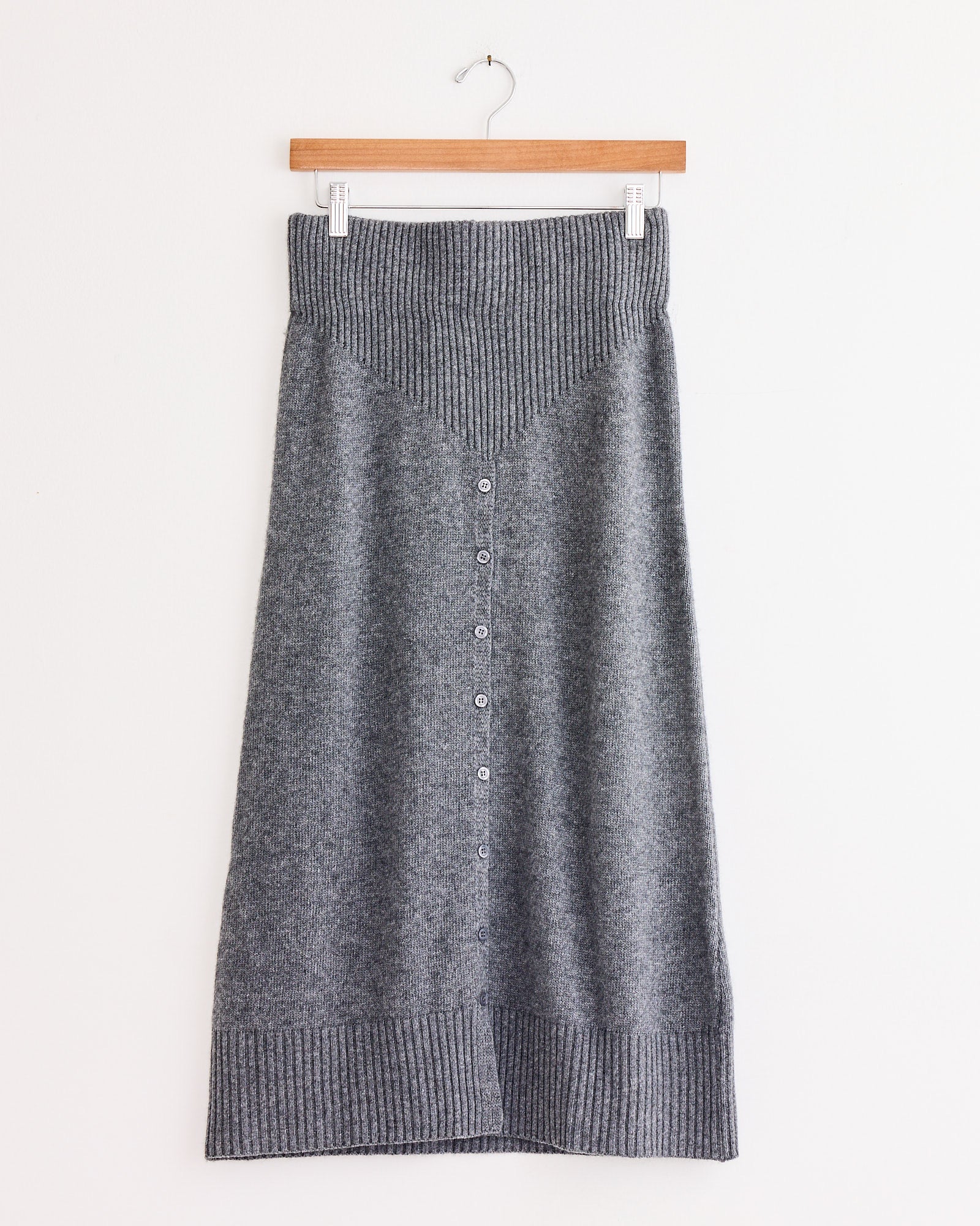 The Amina Skirt in Light Grey by Beaufille is a knee-length slim knit skirt with a ribbed waistband and hem, button-down front, and a V-shaped ribbed detail near the waist, shown on a wooden hanger against a white wall.