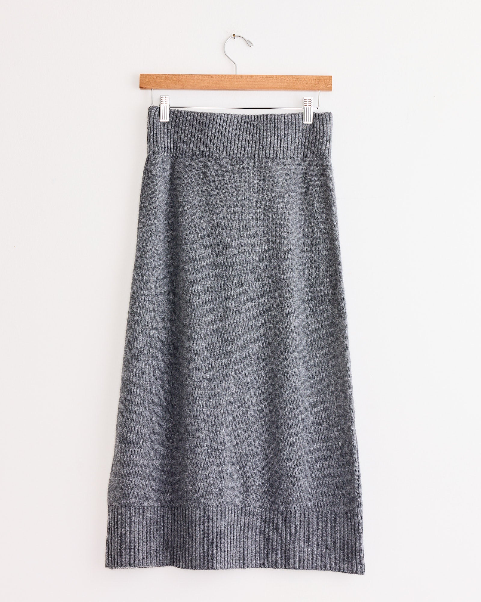 The Amina Skirt in Light Grey by Beaufille is a knee-length slim knit skirt with a ribbed waistband and hem, shown on a wooden hanger against a white background.