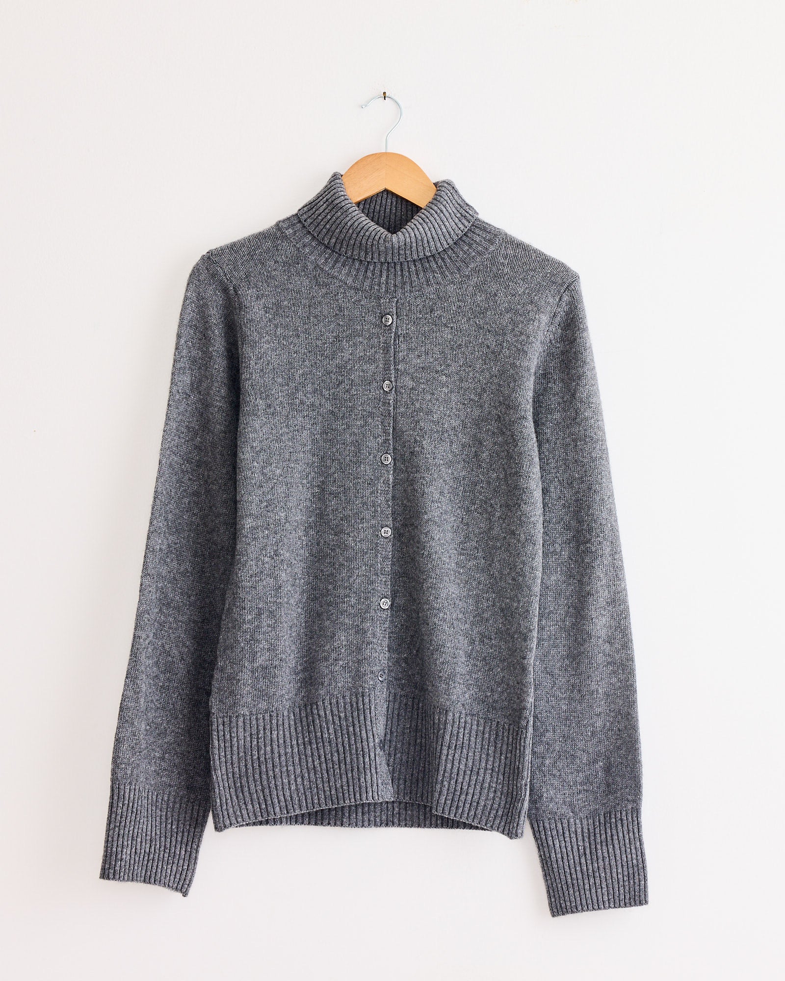 The Edith Sweater in Light Grey by Beaufille features long sleeves, a button-up front, and ribbed turtleneck, cuffs, and hem. Shown on a wooden hanger against a white wall. Chat with us online for fit or sizing questions.
