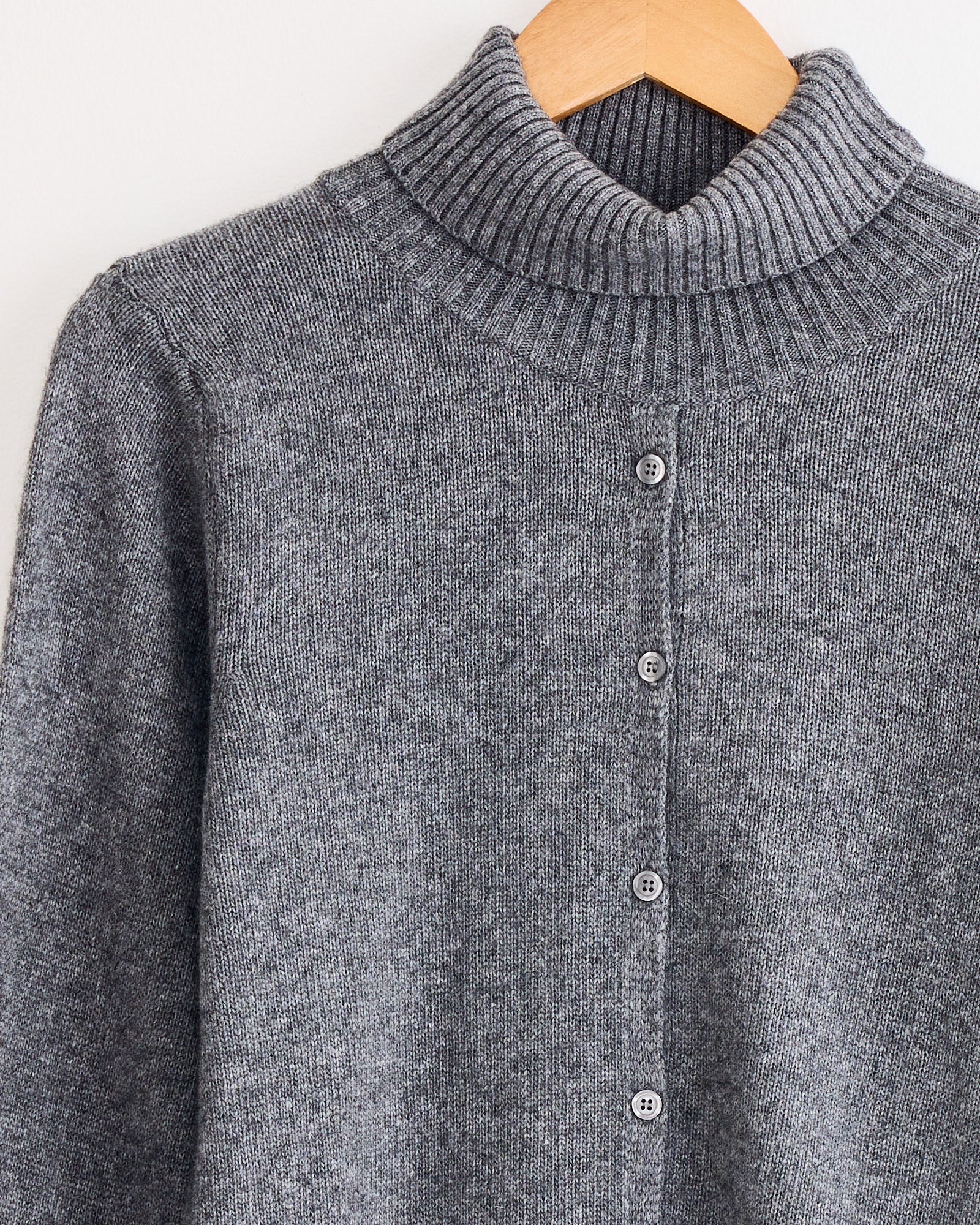 A close-up of the Beaufille Edith Sweater in Light Grey, featuring a ribbed turtleneck and button-down front, displayed on a wooden hanger. For fit or sizing questions, contact us via online chat.