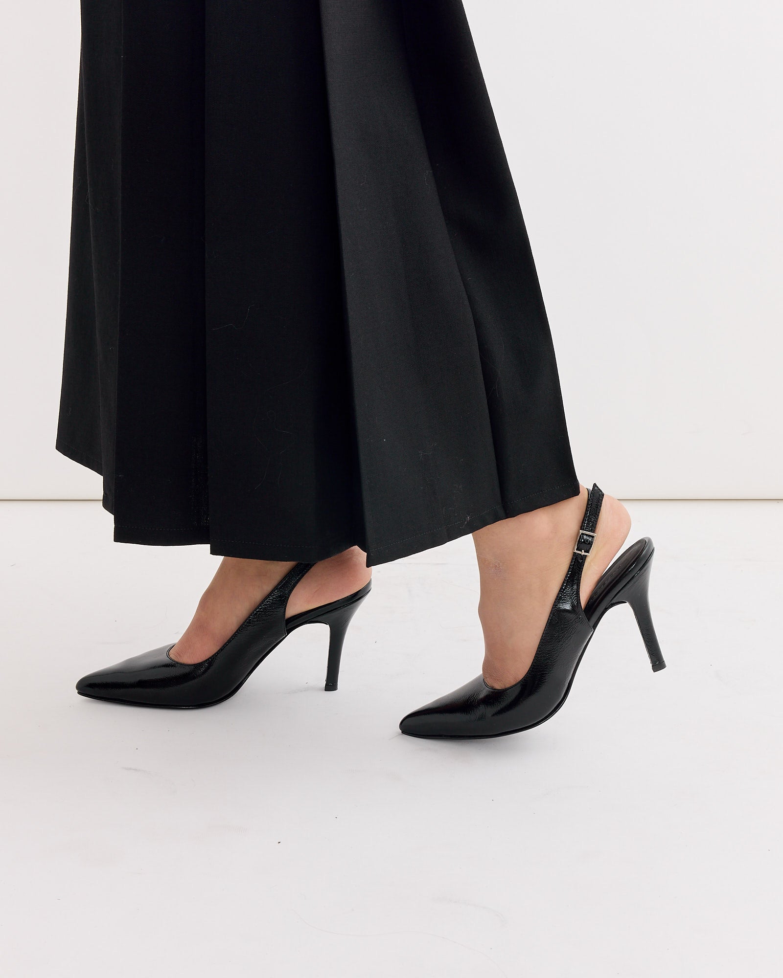 Only the lower legs and feet are visible as a person stands on a white floor, wearing long black pants and the Rachel Comey Cheke Sling Back Heel in Black.