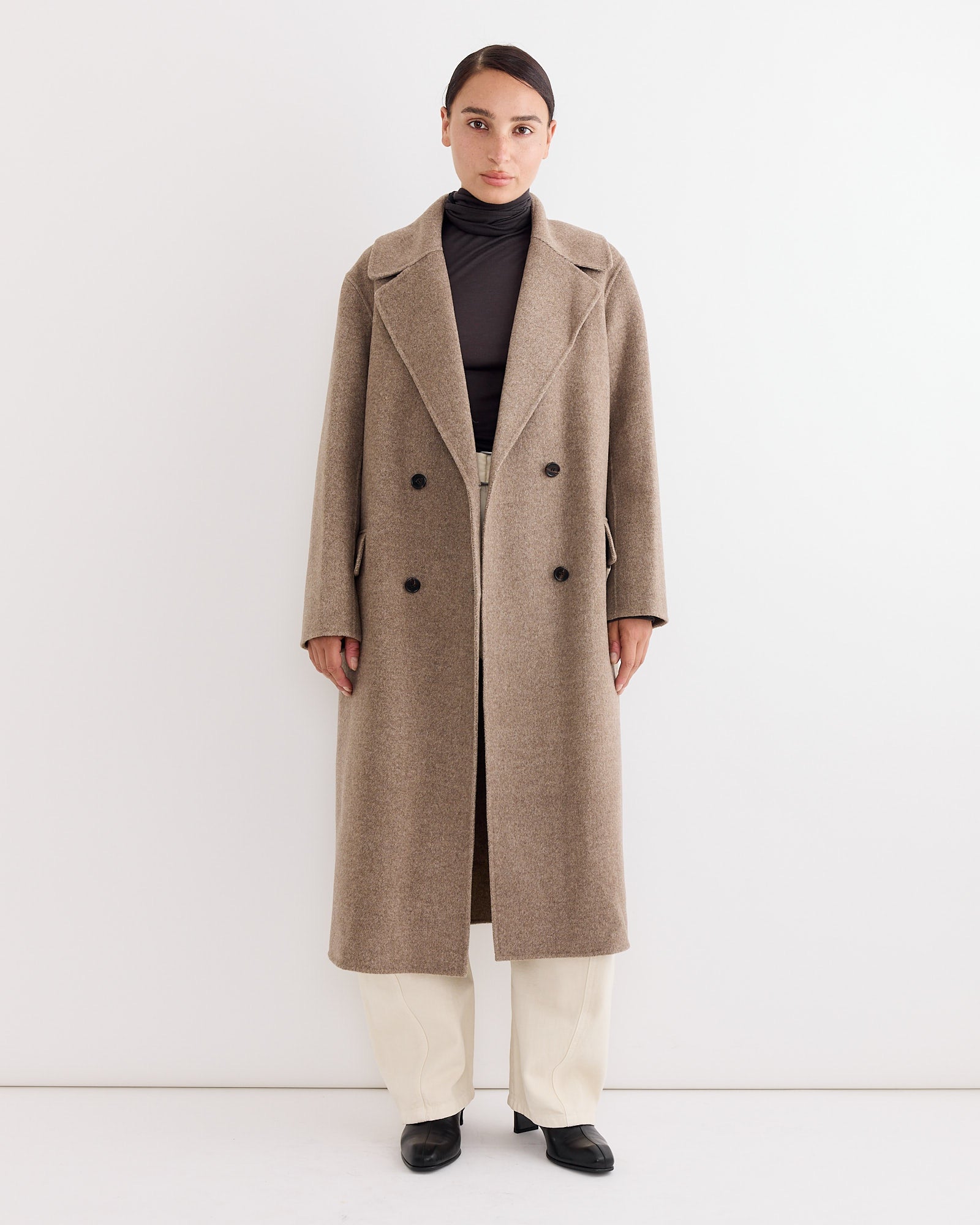 A person stands against a white background, wearing the Rachel Comey Axel Coat in Light Brown over a dark turtleneck, with light pants and black shoes, looking directly at the camera with a neutral expression.