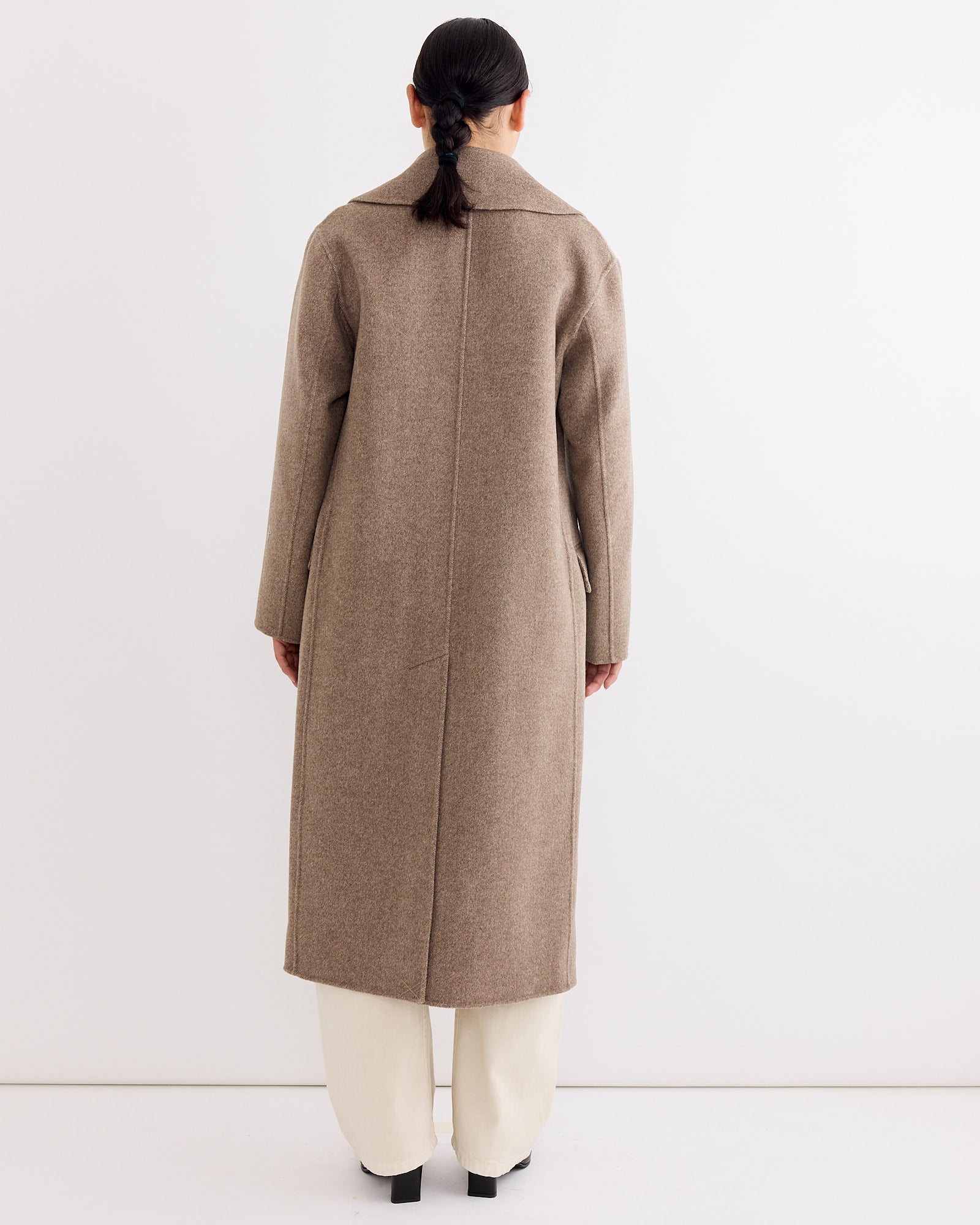 A person with dark hair in a low ponytail is shown from the back against a plain light background, wearing the Rachel Comey Axel Coat in Light Brown and off-white pants.