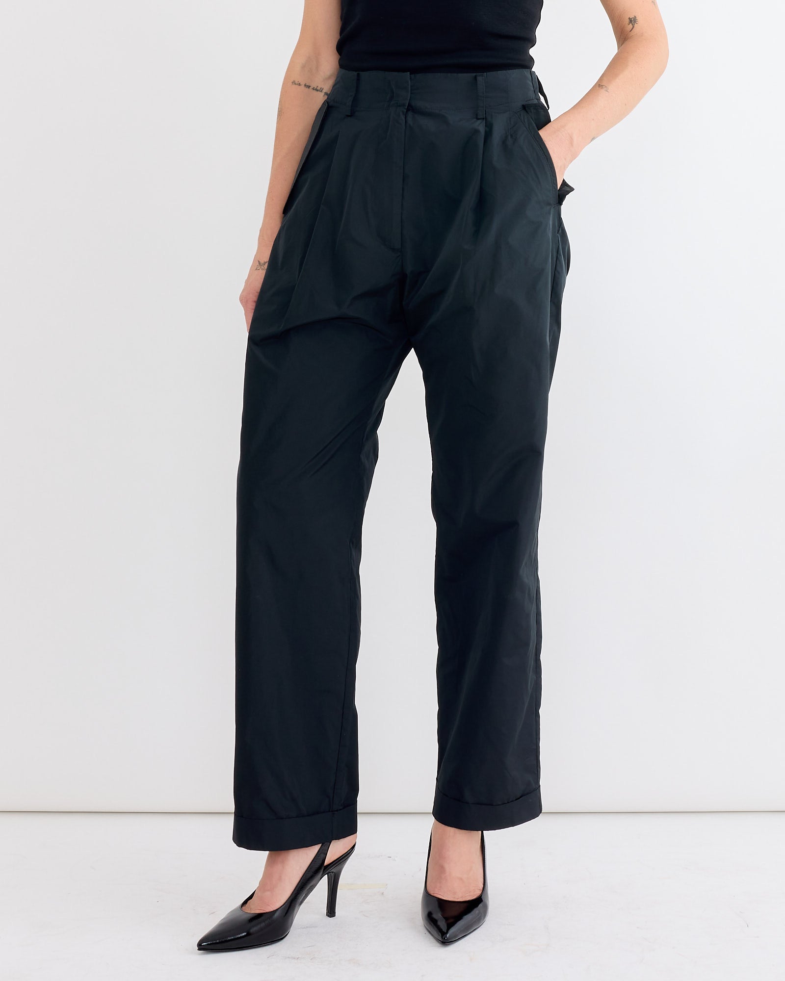 A person models the Rachel Comey Bending Pant in Black—high-waisted, loose-fit recycled polyester—with hands in pockets and black heels, standing against a white background. Only their torso and legs are visible.