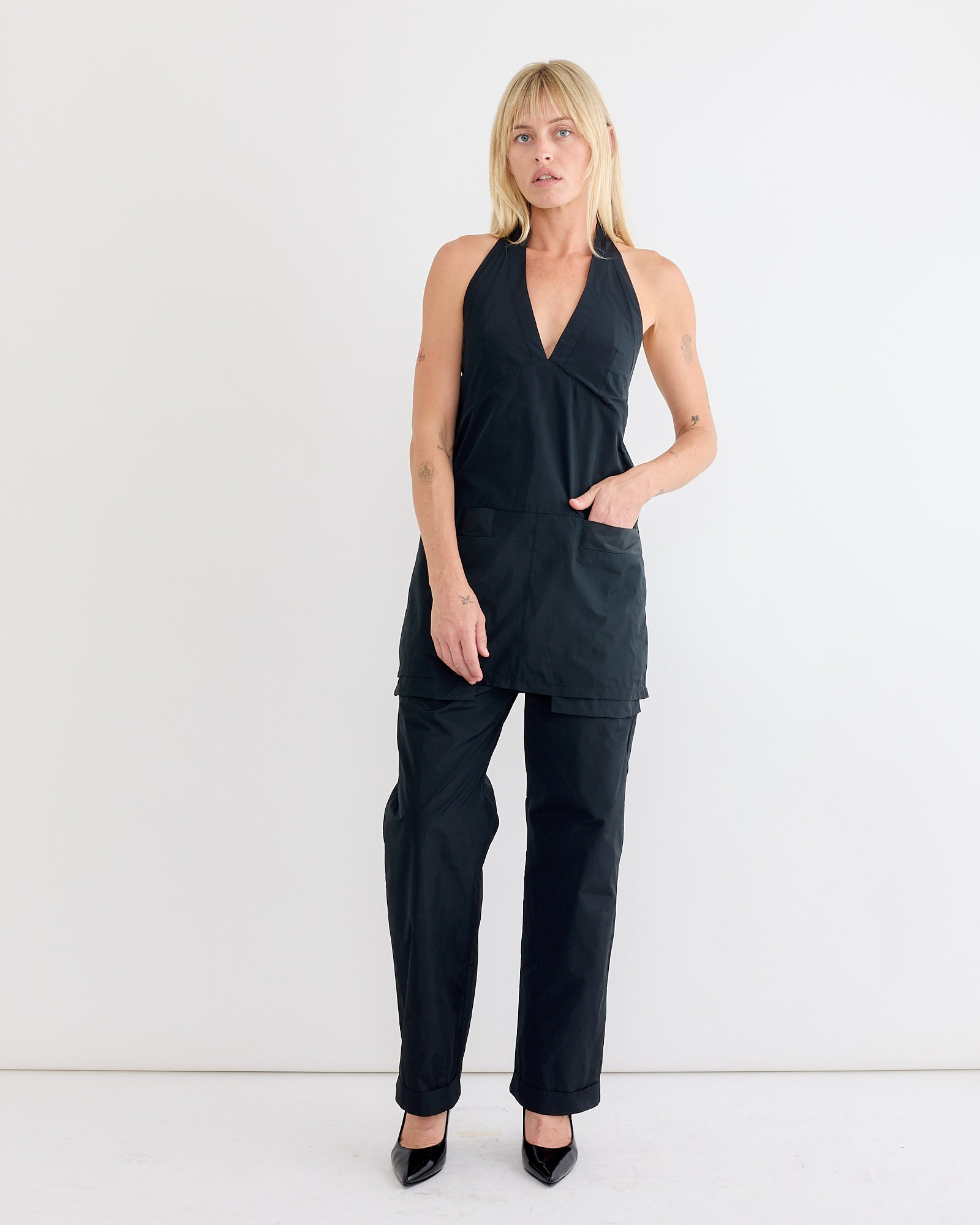 A blonde woman wears the Rachel Comey Onga Dress in Black, a water-resistant taffeta halter-neck style, standing against a plain white background with one hand in her pocket.