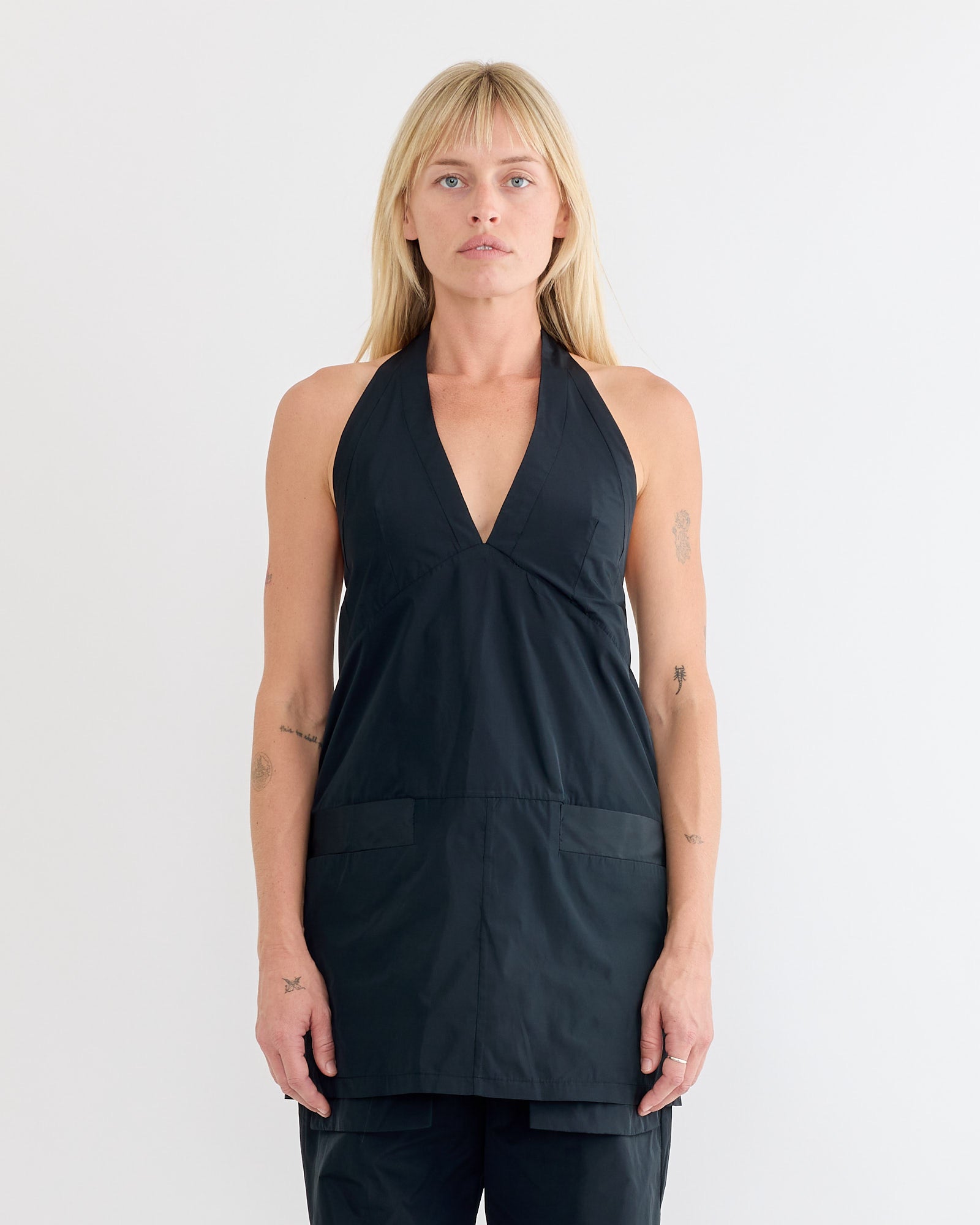 A person with straight blonde hair models the Rachel Comey Onga Dress in Black, a sleeveless deep V-neck halter style, standing against a plain white background and gazing neutrally at the camera.