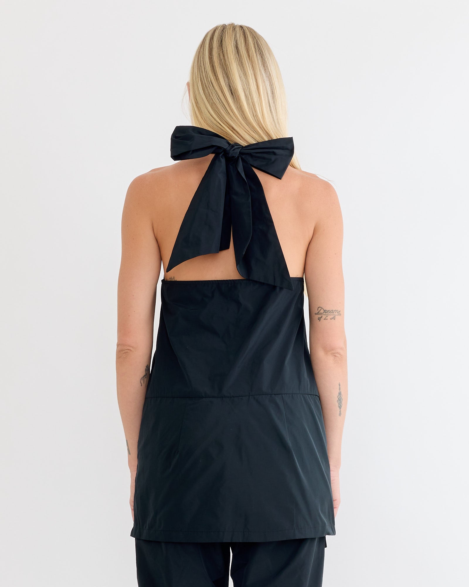 A woman with long blonde hair, seen from behind, wears the Rachel Comey Onga Dress in Black—a sleek, open-back halter-neck dress made from water-resistant taffeta and styled with matching black bottoms.