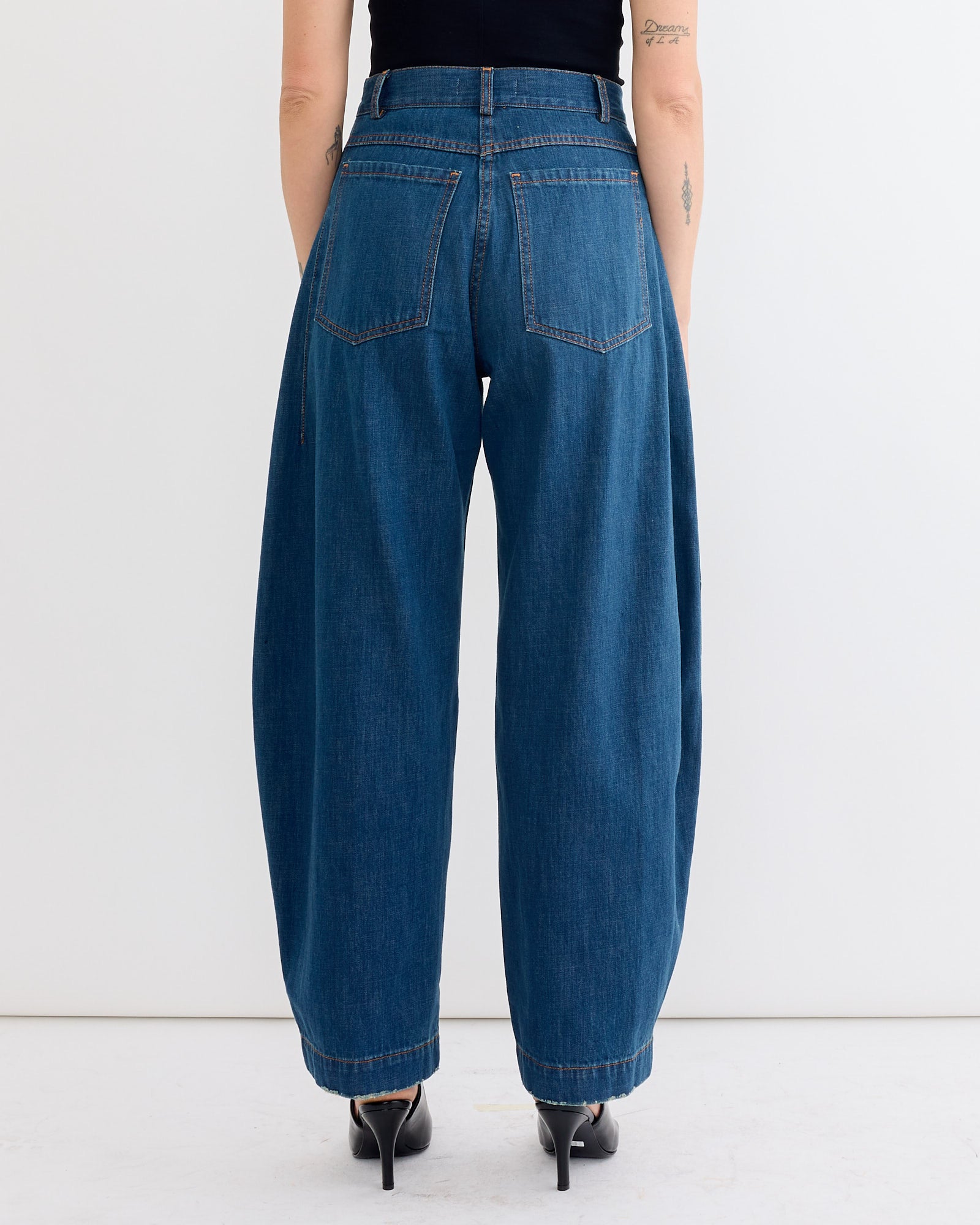 Person shown from behind, wearing the Rachel Comey Keenan Pant in Vintage Indigo—loose-fitting, wide-leg jeans with large back pockets—paired with a black top and black heeled shoes against a white background.