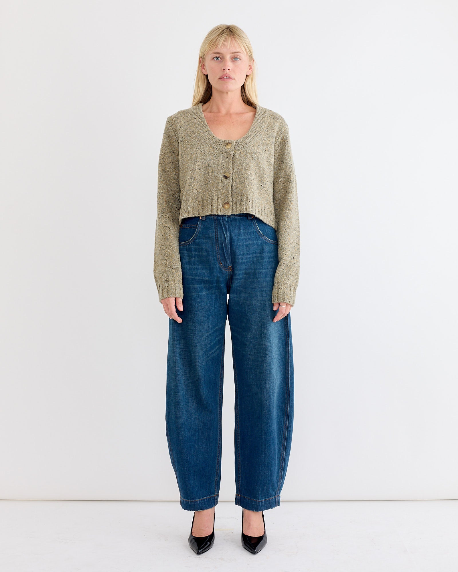 A person stands against a white background wearing the Rachel Comey Uridge Cardigan in Stone, paired with wide-leg blue jeans and black heels. They have straight blonde hair and a neutral expression.