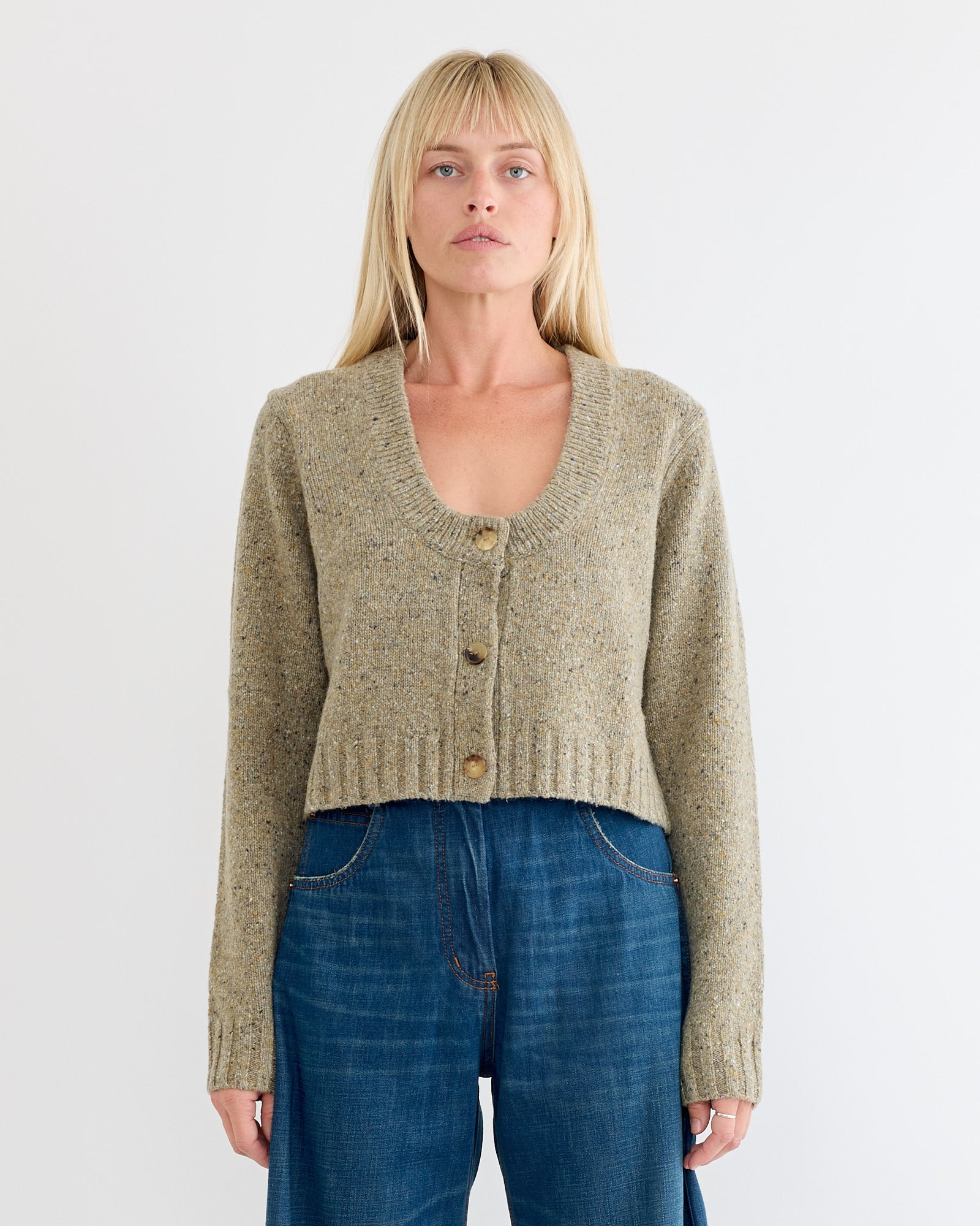 A woman with straight blonde hair wears the Rachel Comey Uridge Cardigan in Stone and high-waisted blue jeans, standing against a plain white background.