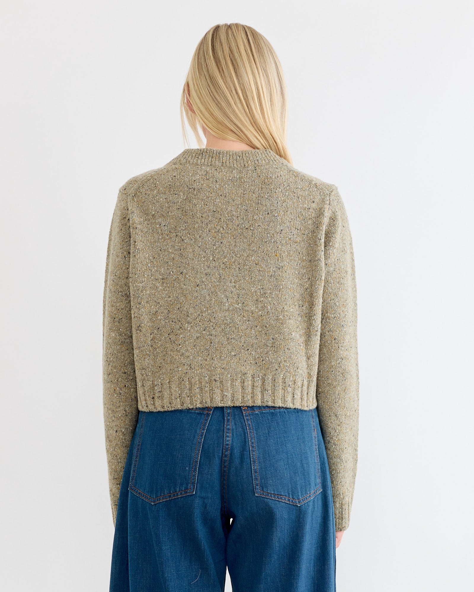 A woman with long blonde hair, seen from behind, wears the Rachel Comey Uridge Cardigan in Stone and high-waisted blue jeans against a plain white background.