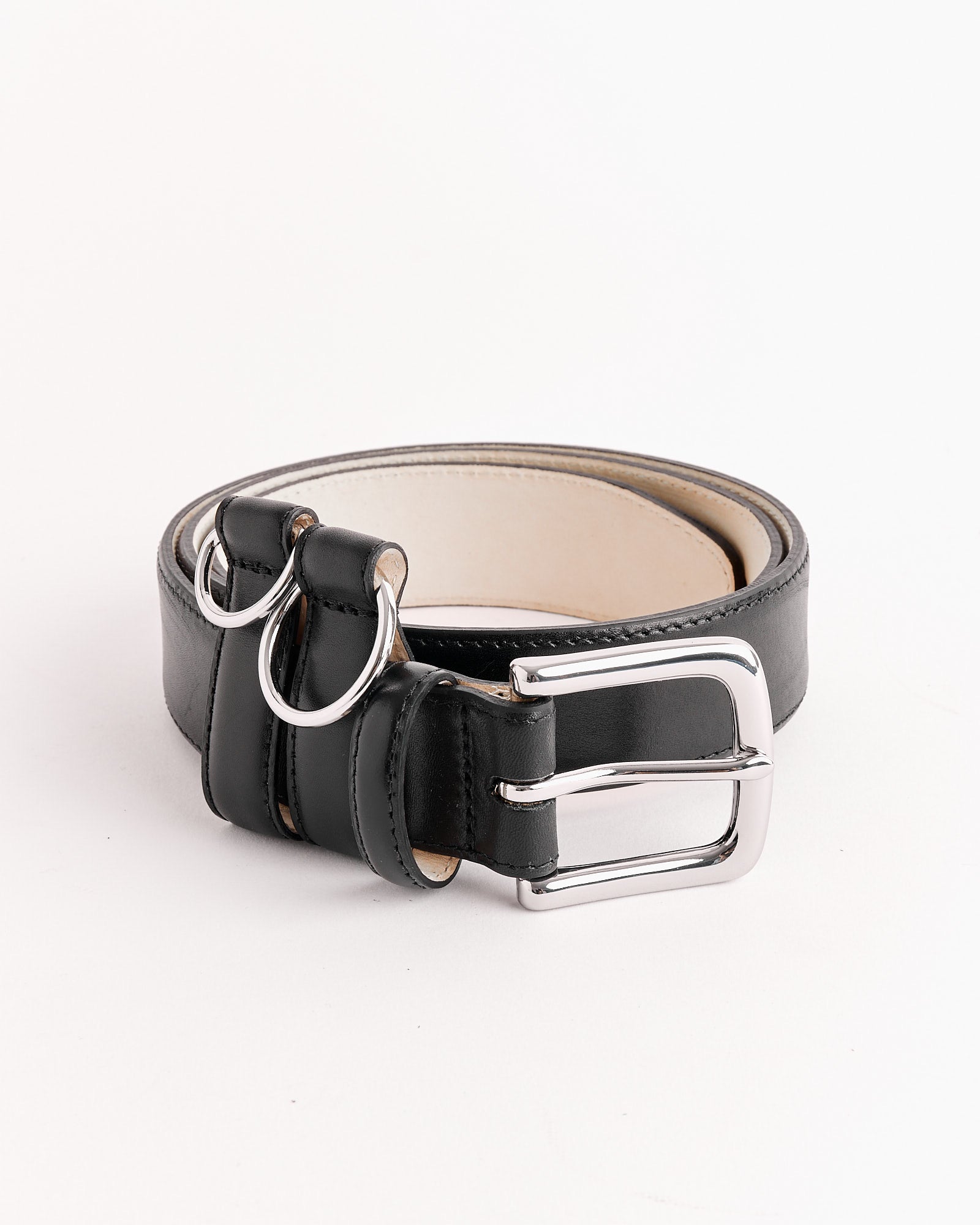 The Tibi Classic Ring Hardware Leather Belt in Black features a silver buckle, two silver D-rings, and a light inner lining. Shown coiled on a white surface. For fit or sizing questions, contact our online chat team.