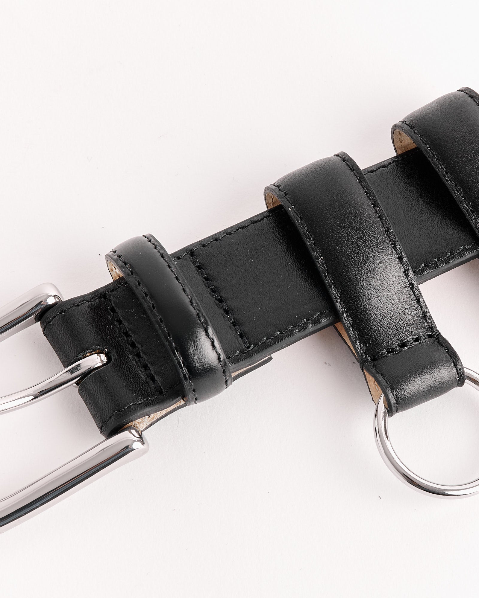 Close-up of the Tibi Classic Ring Hardware Leather Belt in Black, featuring a silver metal buckle and loops on a white background. For sizing or fit questions, connect with our online chat team.