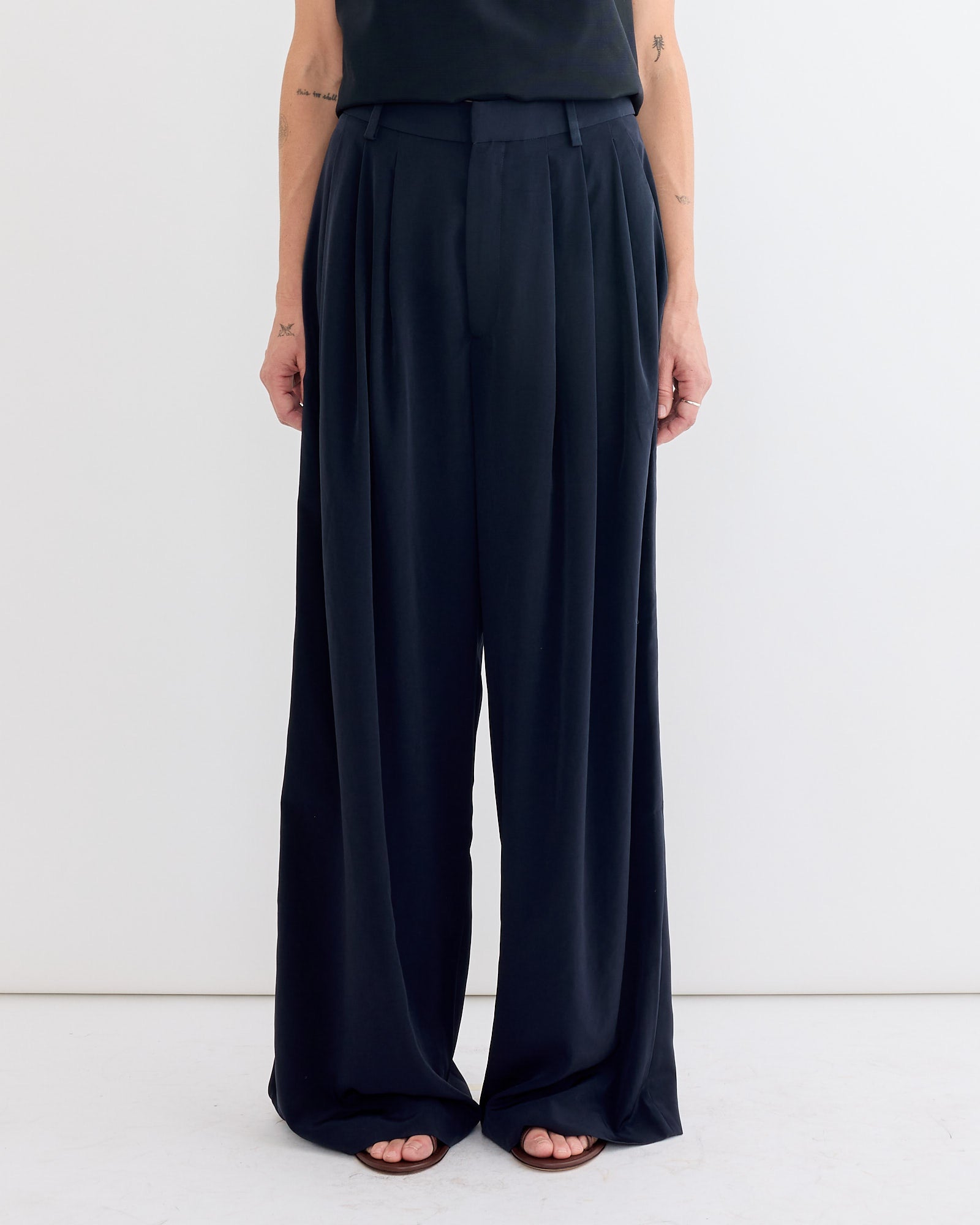 A person models Tibi's Relaxed Suiting Triple Pleat Pant in Navy, featuring a wide-leg, high-waisted silhouette, paired with a black top, standing against a white background. Only their lower torso and legs are visible.