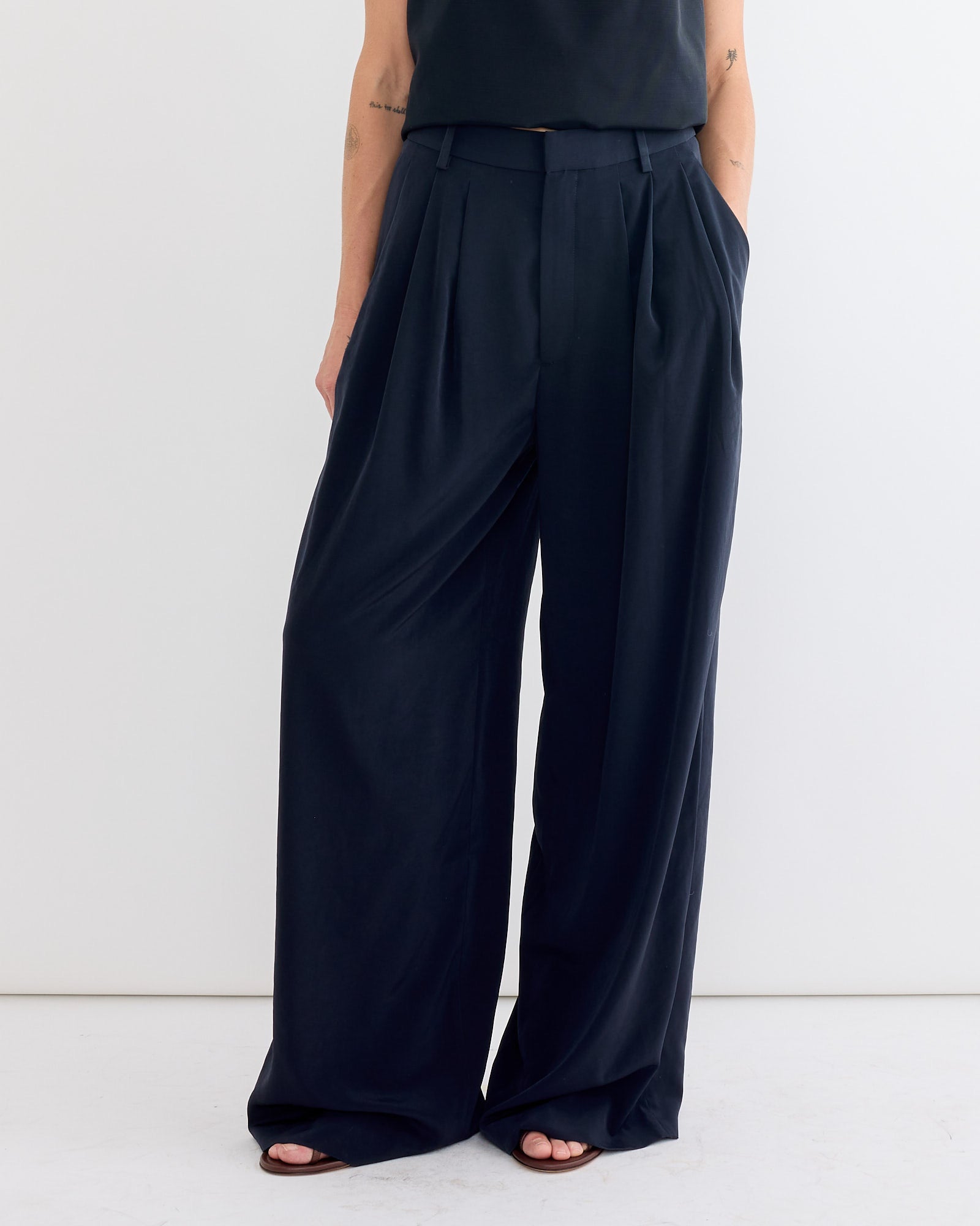 A person models the Tibi Relaxed Suiting Triple Pleat Pant in Navy, standing against a plain white background with hands in pockets. Only the lower torso and legs are shown, highlighting the high-waisted, wide-leg design.