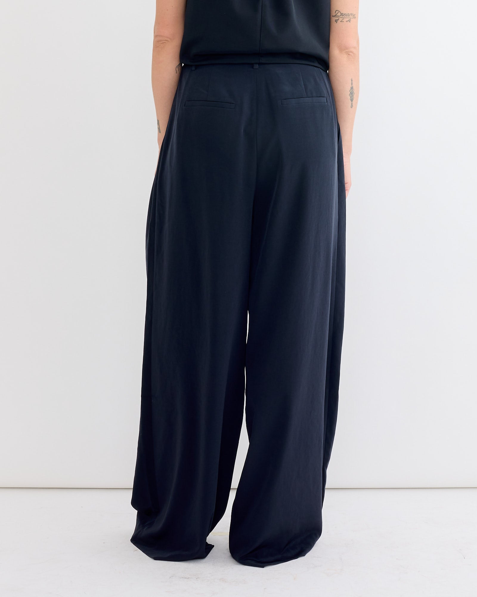 A person stands with their back to the camera, highlighting the Tibi Relaxed Suiting Triple Pleat Pant in Navy. Only the lower back and wide-leg silhouette are visible against a plain white background.