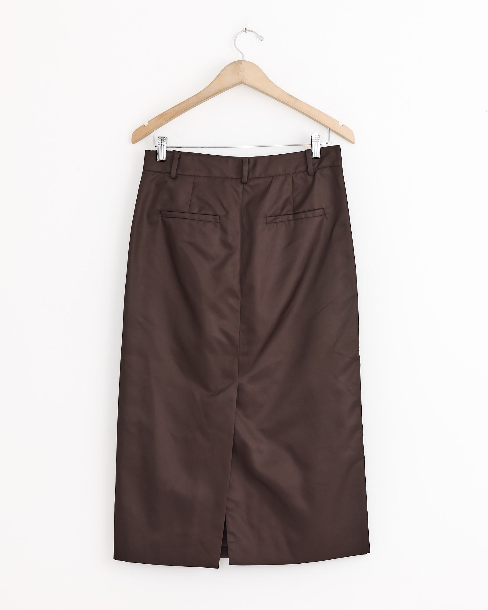 The Tibi Eco Nylon Midi Trouser in Skirt Brown is a knee-length, utility-inspired skirt with a front slit and two back welt pockets, shown on a wooden hanger against a plain white background.