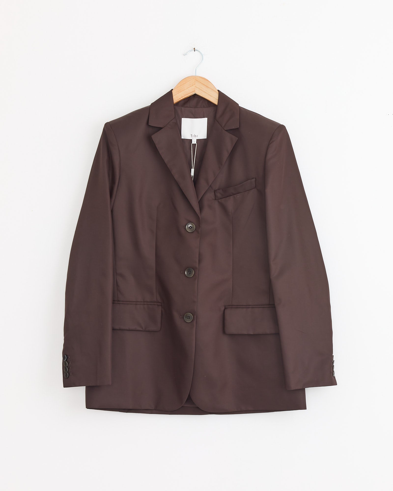 The Tibi Eco Nylon Max Blazer in Brown, featuring a water-repellent finish, three buttons, notched lapel, and front pockets, hangs on a wooden hanger against a plain white background.