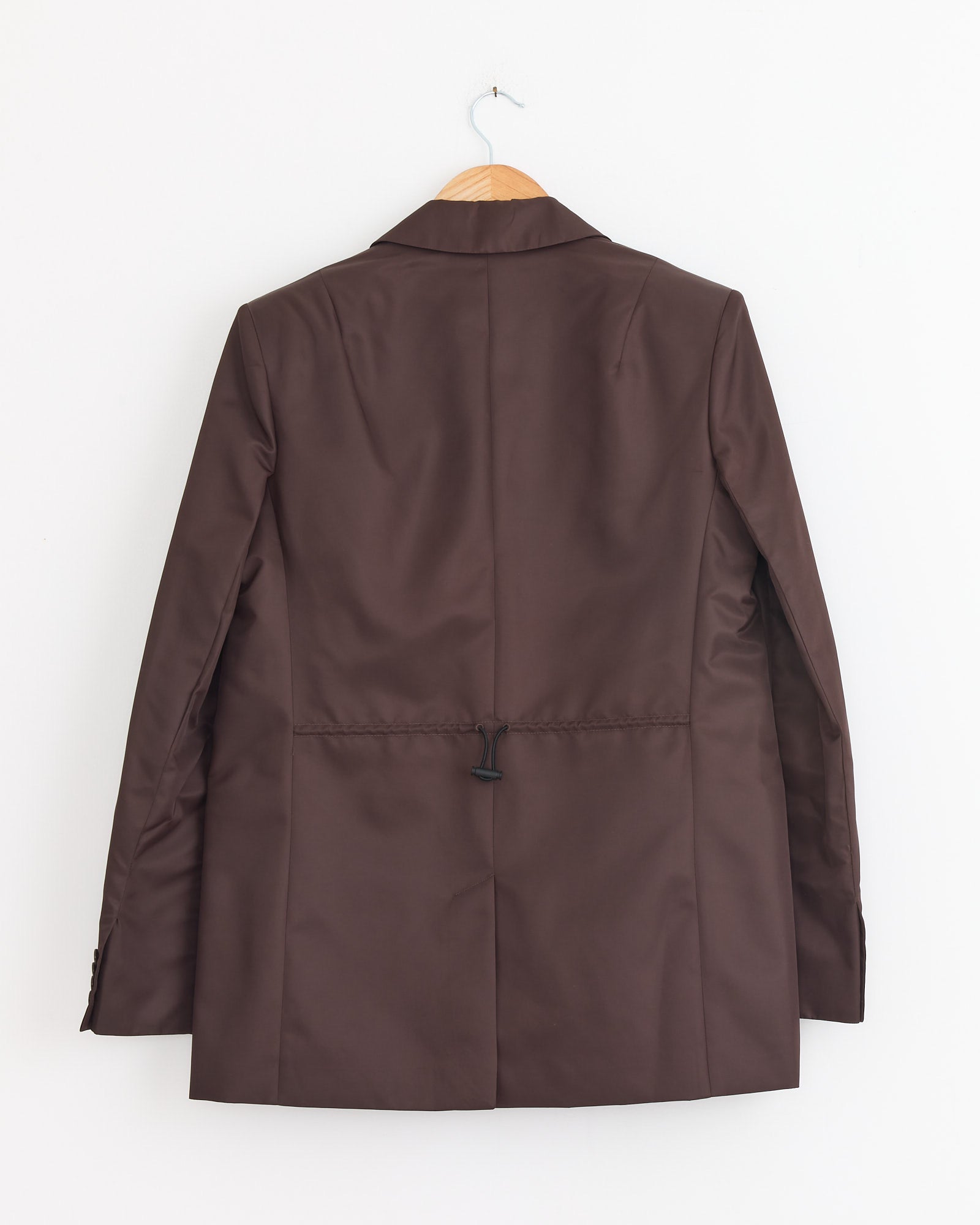 The Tibi Eco Nylon Max Blazer in Brown, featuring a water-repellent finish, hangs on a wooden hanger with its back facing forward against a plain white wall.