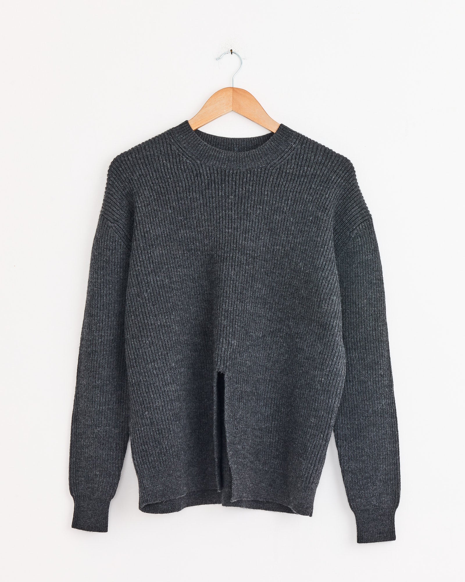 The Tibi Split Front Easy Pullover in Dark Heather Grey, a wool knit sweater with long sleeves and a crew neckline, hangs on a wooden hanger. Its modern design features a vertical slit at the front hem.