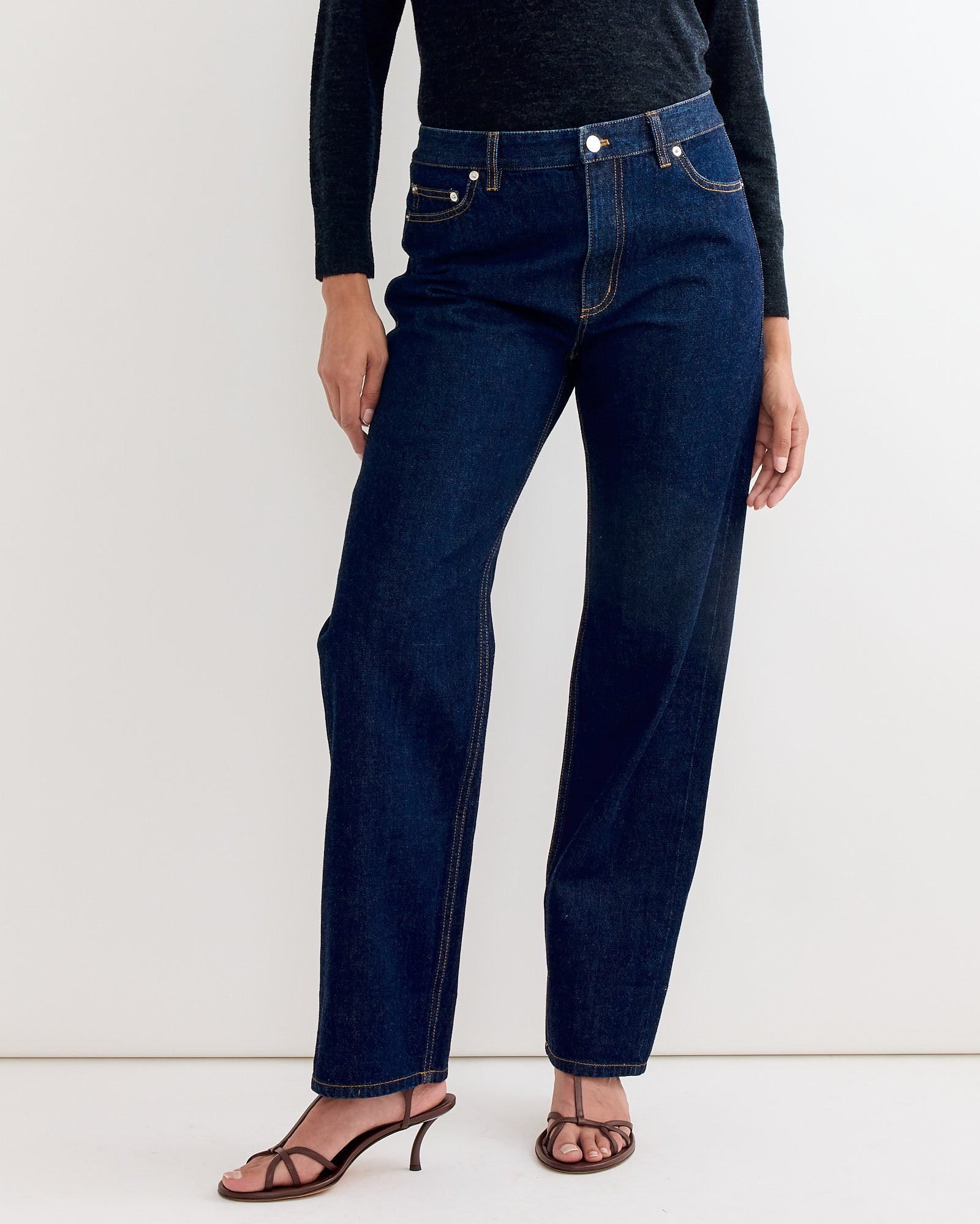 A person wears the Tibi 5 Pocket Alec Jean in Dark Denim, styled slouchy with a dark long-sleeve top and brown heeled sandals, standing against a plain white background.