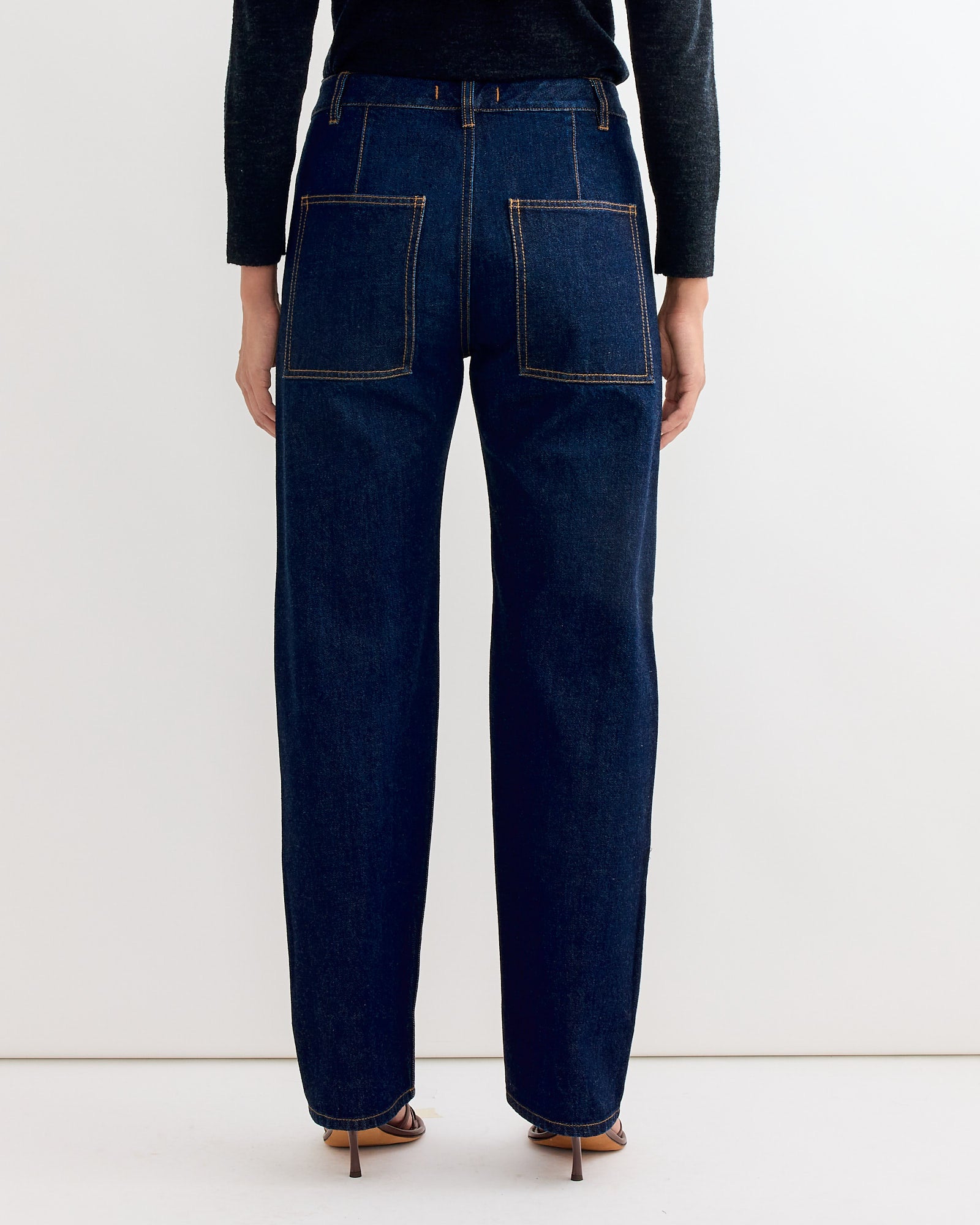 A person is seen from the back wearing Tibi’s 5 Pocket Alec Jean in Dark Denim—featuring a slouchy fit, large square back pockets, and tan stitching—styled with brown heeled sandals and a black long-sleeve top.