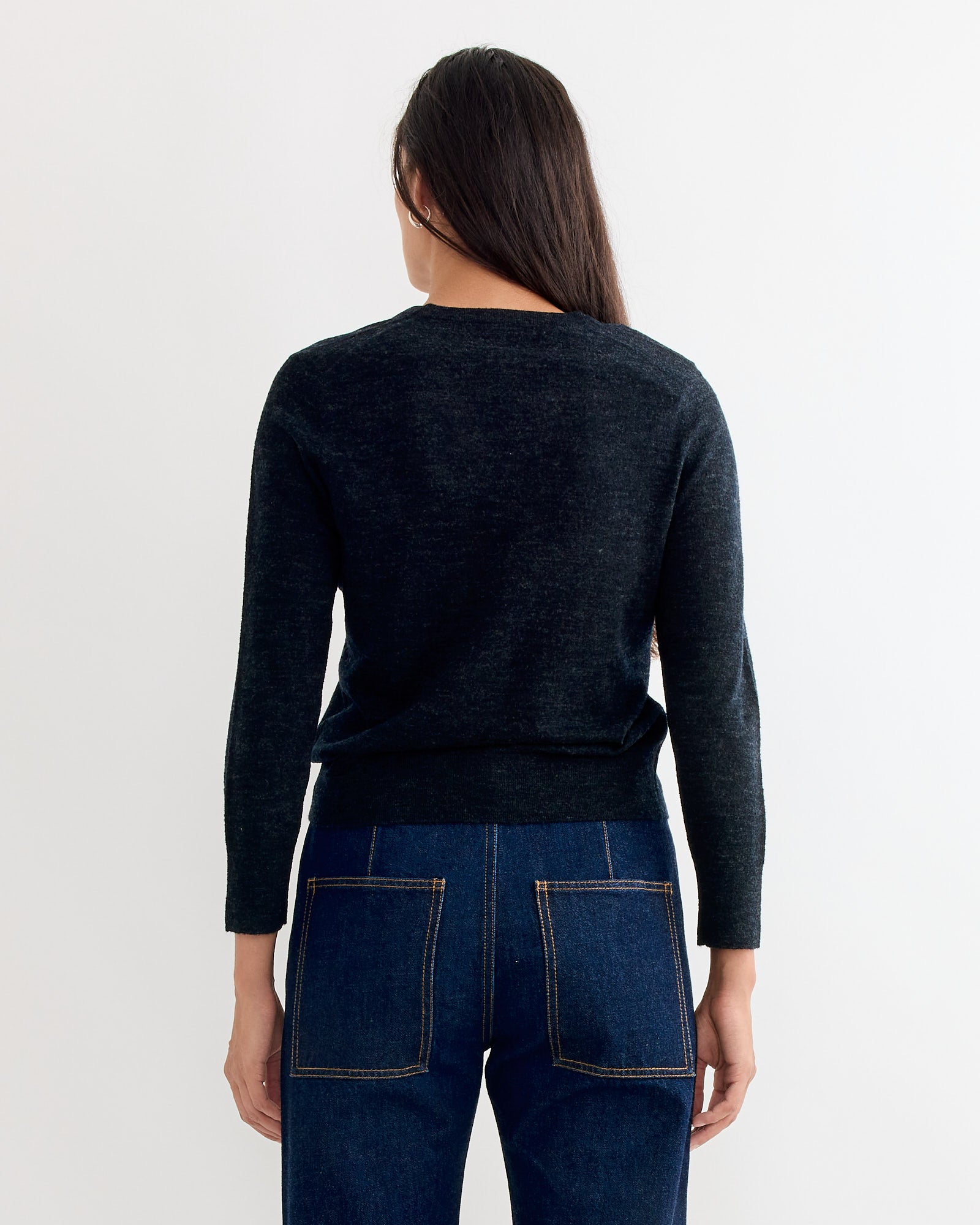 A person with long dark hair stands facing away, wearing Tibi’s Wool Shrunken V Neck Sweater in Charcoal and high-waisted jeans with large back pockets, set against a plain light background.