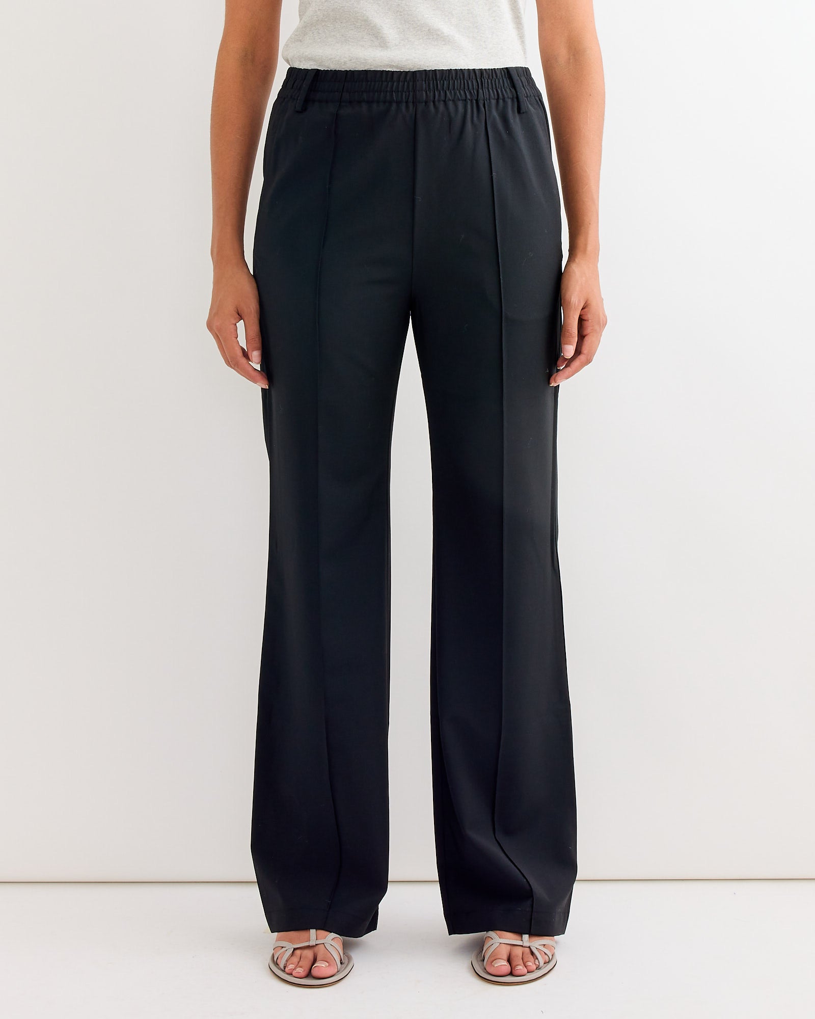 Only the lower half is shown: a person stands against a white wall, wearing Tibi’s Tropical Wool Scottie Jogger in Black—wide-leg, high-waisted pants with an elastic waistband—paired with a light top and open-toe sandals.