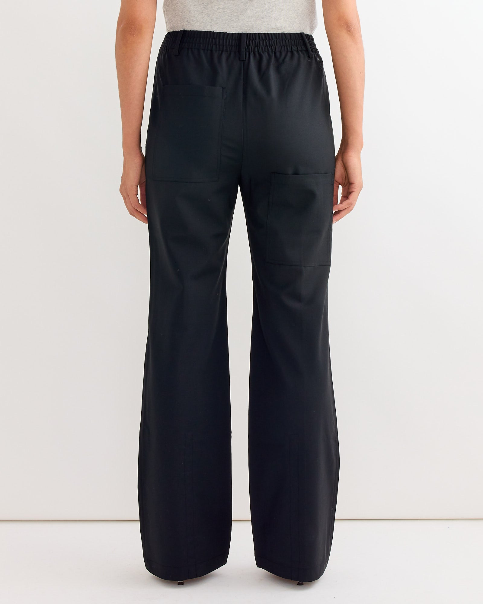 A person stands facing away, wearing Tibis Tropical Wool Scottie Jogger in Black, featuring an elastic waistband and two large back pockets, paired with a light gray top against a plain white background.