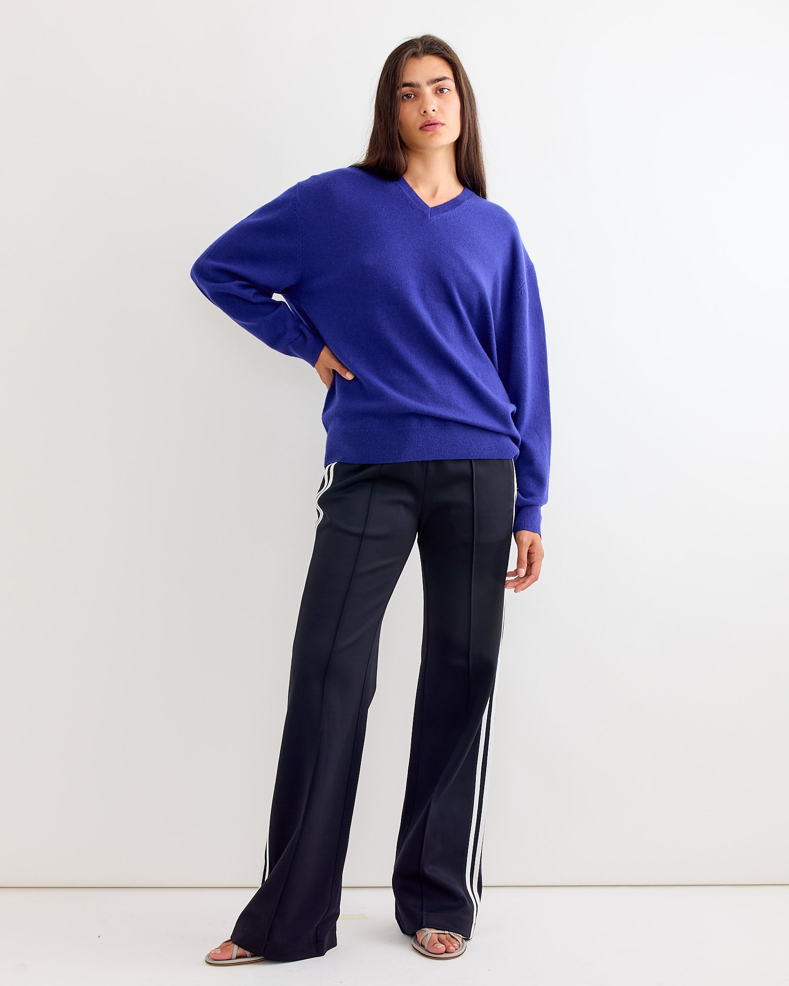 A person with long dark hair stands against a white background, wearing a loose cobalt blue sweater and Tibi’s Active Knit Scottie Striped Jogger in Black Multi with white side stripes, paired with open-toe sandals.