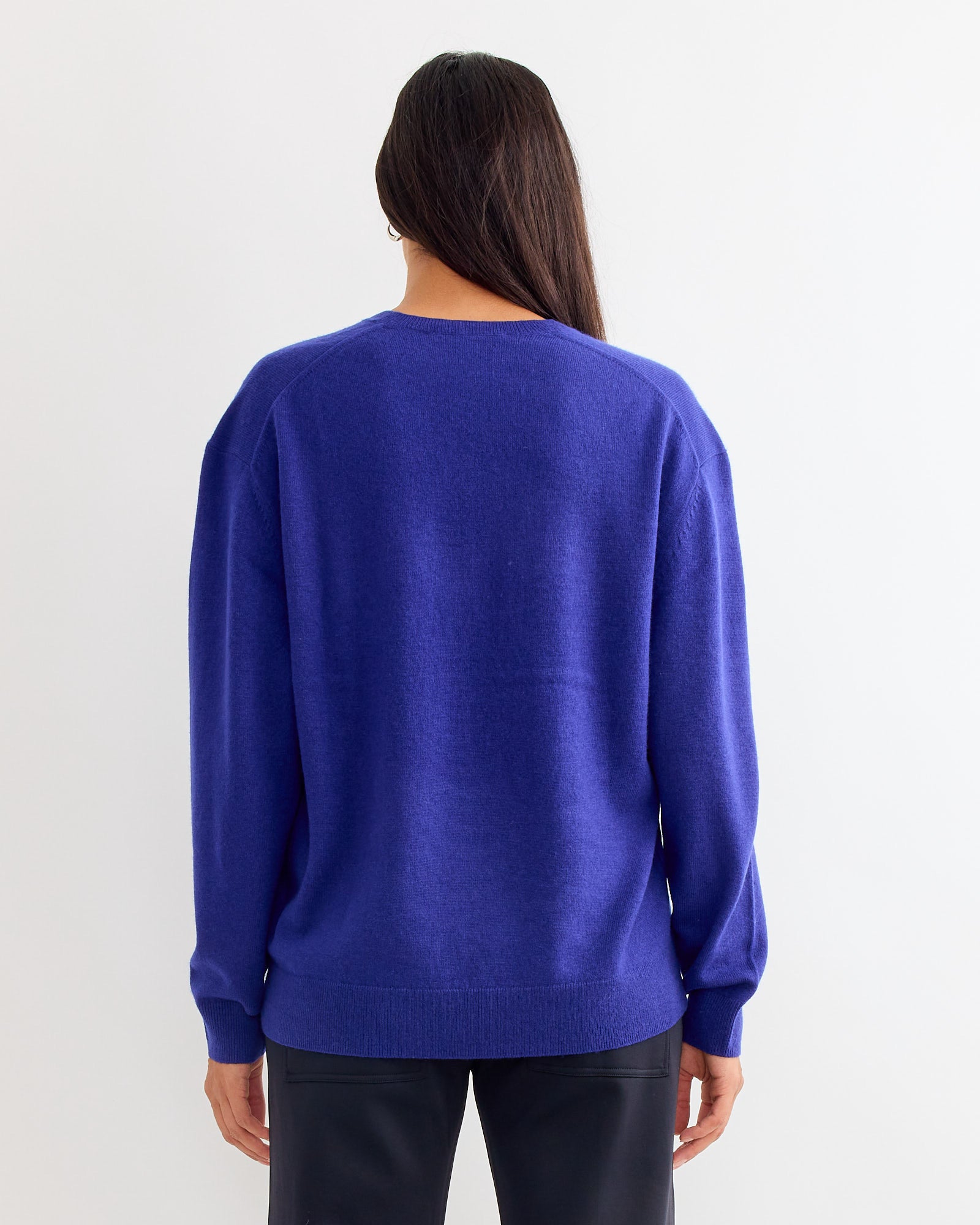 A person with long dark hair, seen from behind, wears Tibis Washable Cashmere V Neck Sweater in Ink Blue and dark pants—perfect for exploring sizing and fit via online chat, all set against a plain white background.