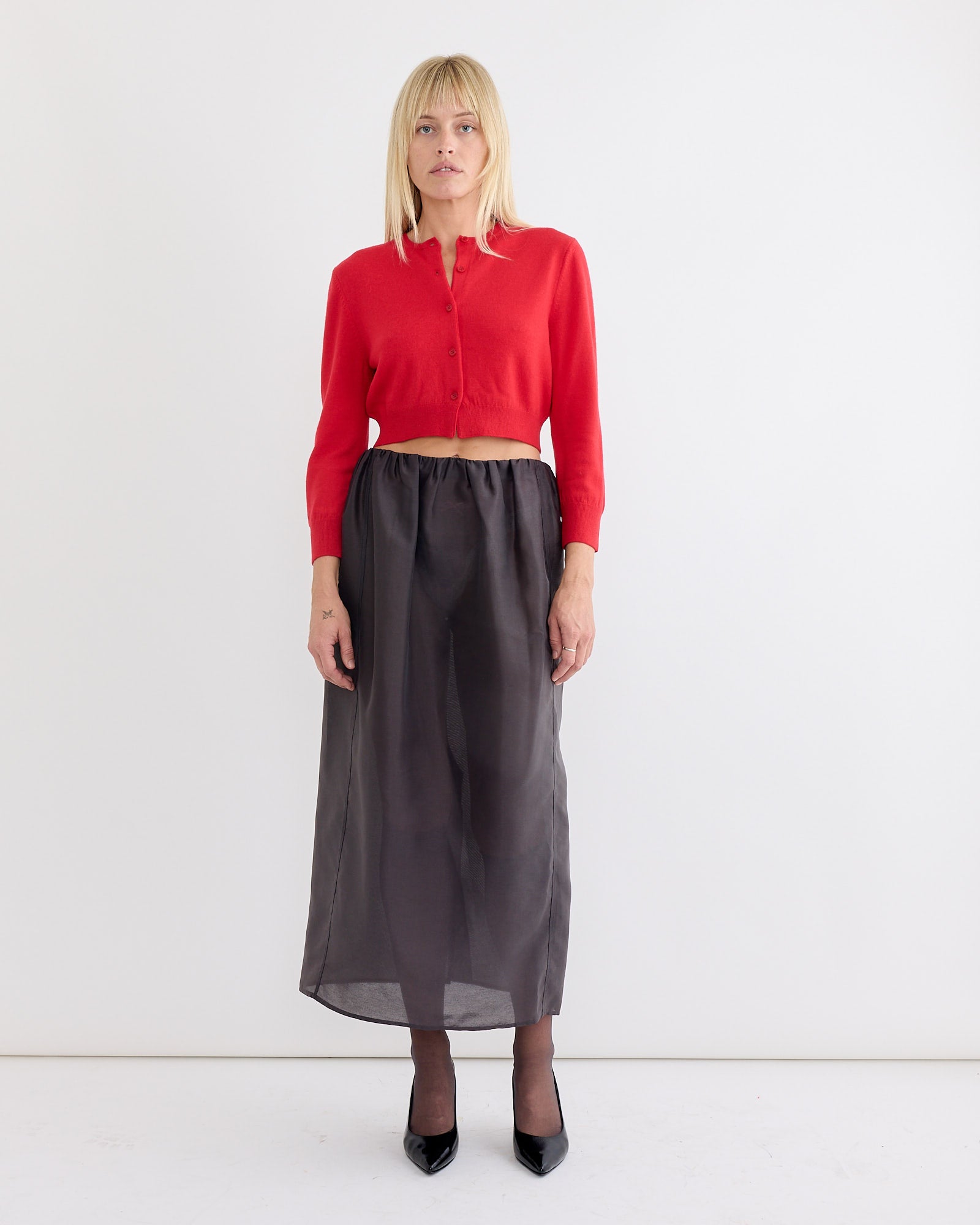 A woman with blonde hair and a neutral expression stands against a white background, wearing Amomento’s Round Neck Crop Cardigan in Red, a high-waisted sheer black maxi skirt, black tights, and black pointed-toe heels.