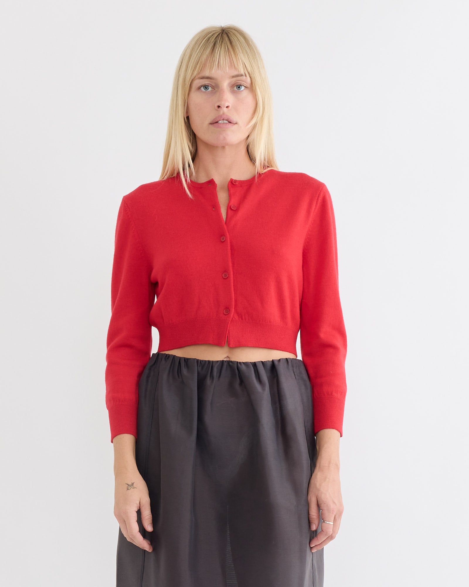 A woman with straight blonde hair wears the Amomento Round Neck Crop Cardigan in Red and a high-waisted dark gray skirt, standing against a plain white background.